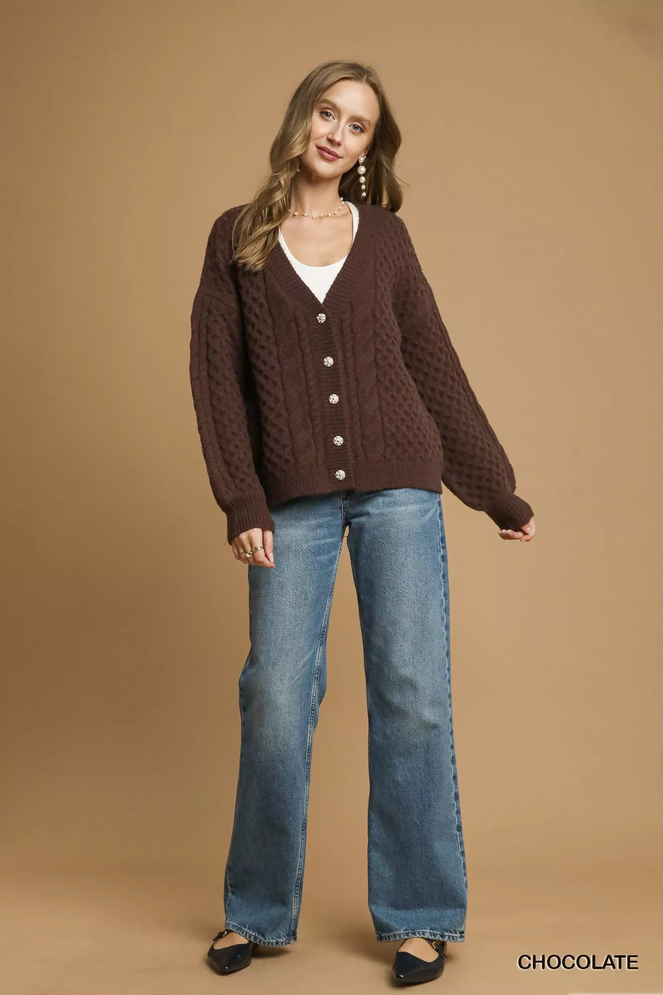 Model wearing a chocolate-brown cable-knit cardigan with pearl buttons, styled with a white tank top and jeans.