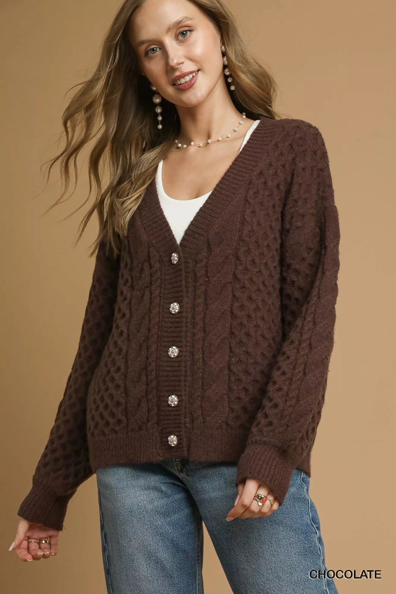 Model wearing a chocolate-brown cable-knit cardigan with pearl buttons, styled with a white tank top and jeans.