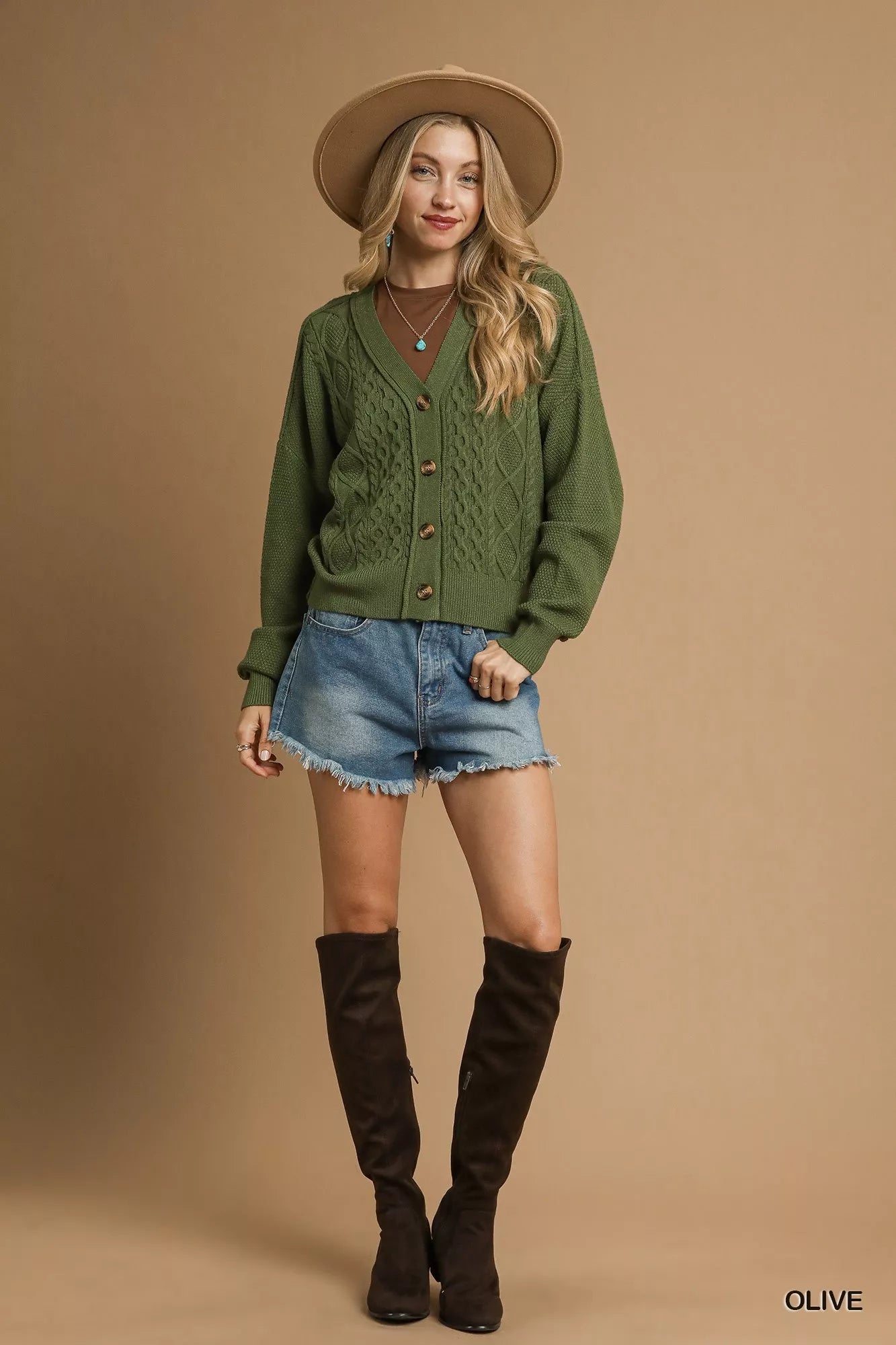 Model wearing an olive green chunky cable knit V-neck cardigan with button-up front, styled with denim shorts and a wide-brimmed hat.