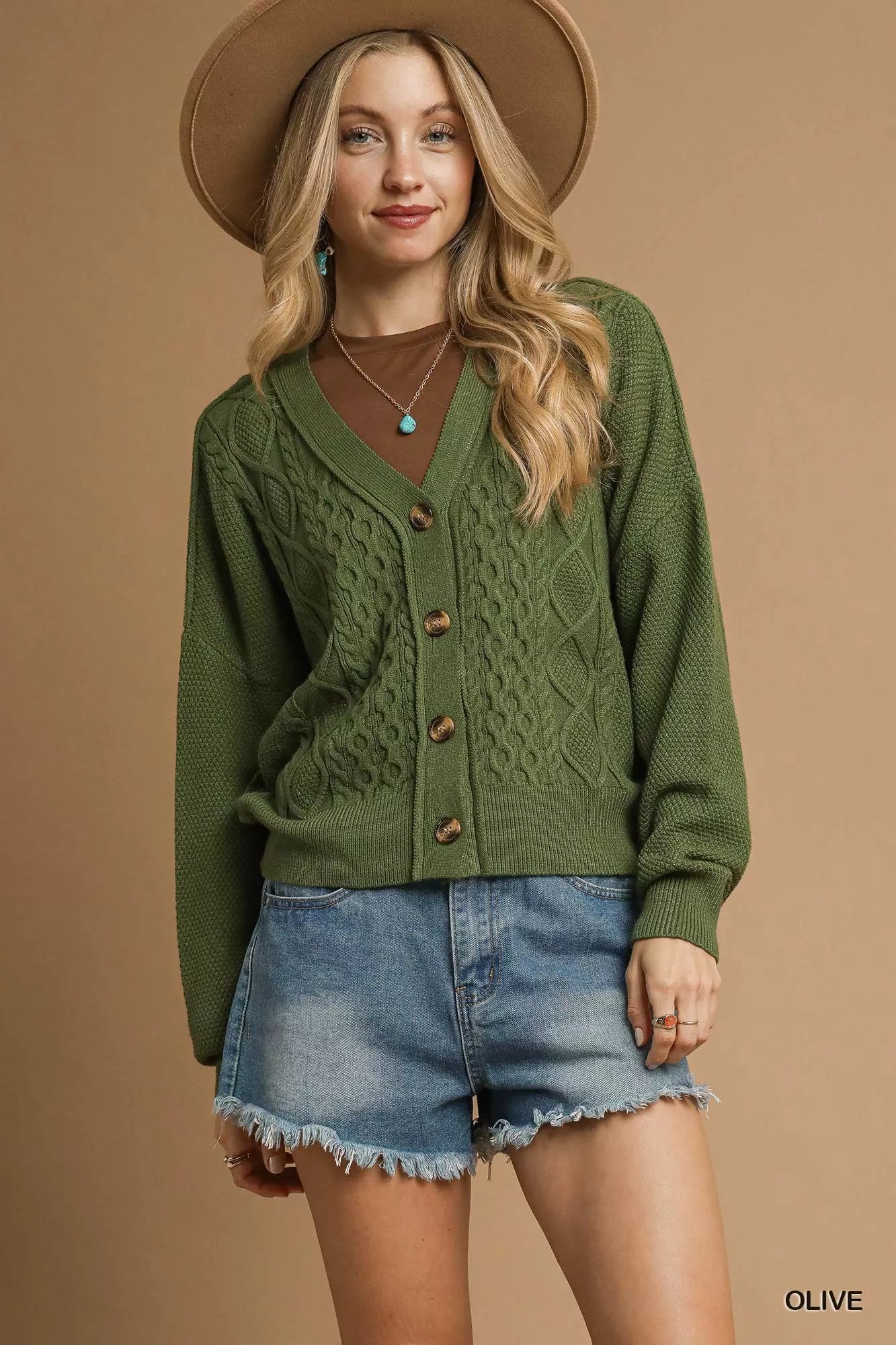 Model wearing an olive green chunky cable knit V-neck cardigan with button-up front, styled with denim shorts and a wide-brimmed hat.