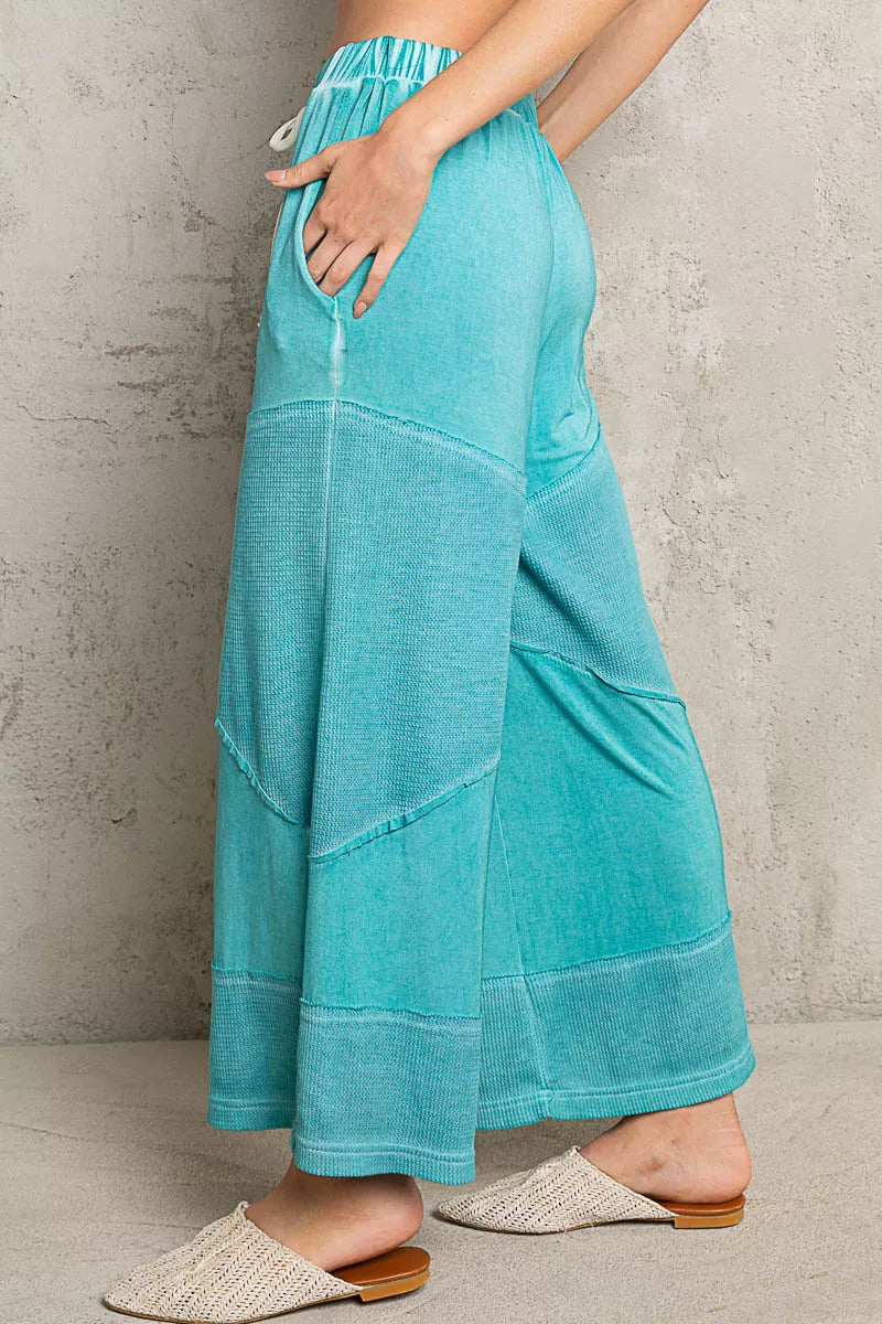 Model wearing teal ribbed wide-leg culotte pants with drawstring waist and textured contrast panels, styled casually with woven flats.