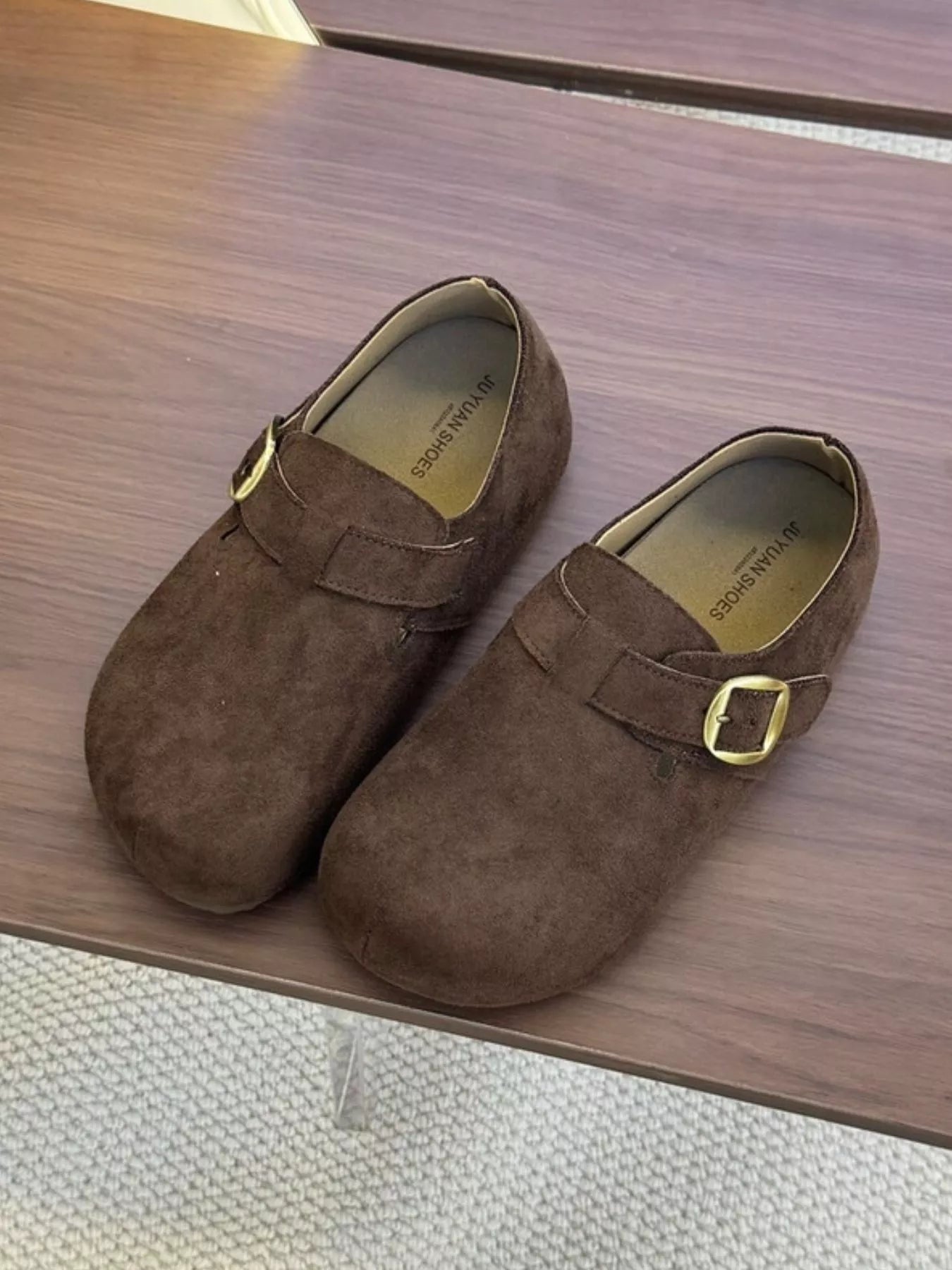 Pair of brown suede buckle clogs with a flat sole displayed on a wooden surface, showcasing the minimalist slip-on design.