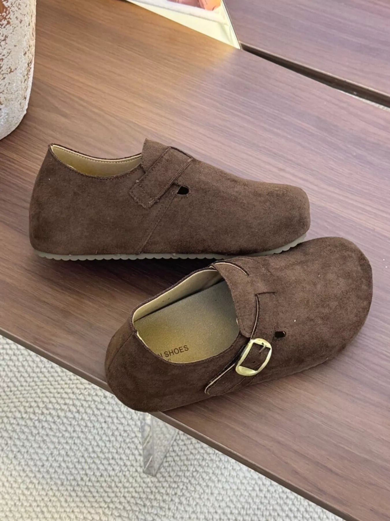 Pair of brown suede buckle clogs with a flat sole displayed on a wooden surface, showcasing the minimalist slip-on design.