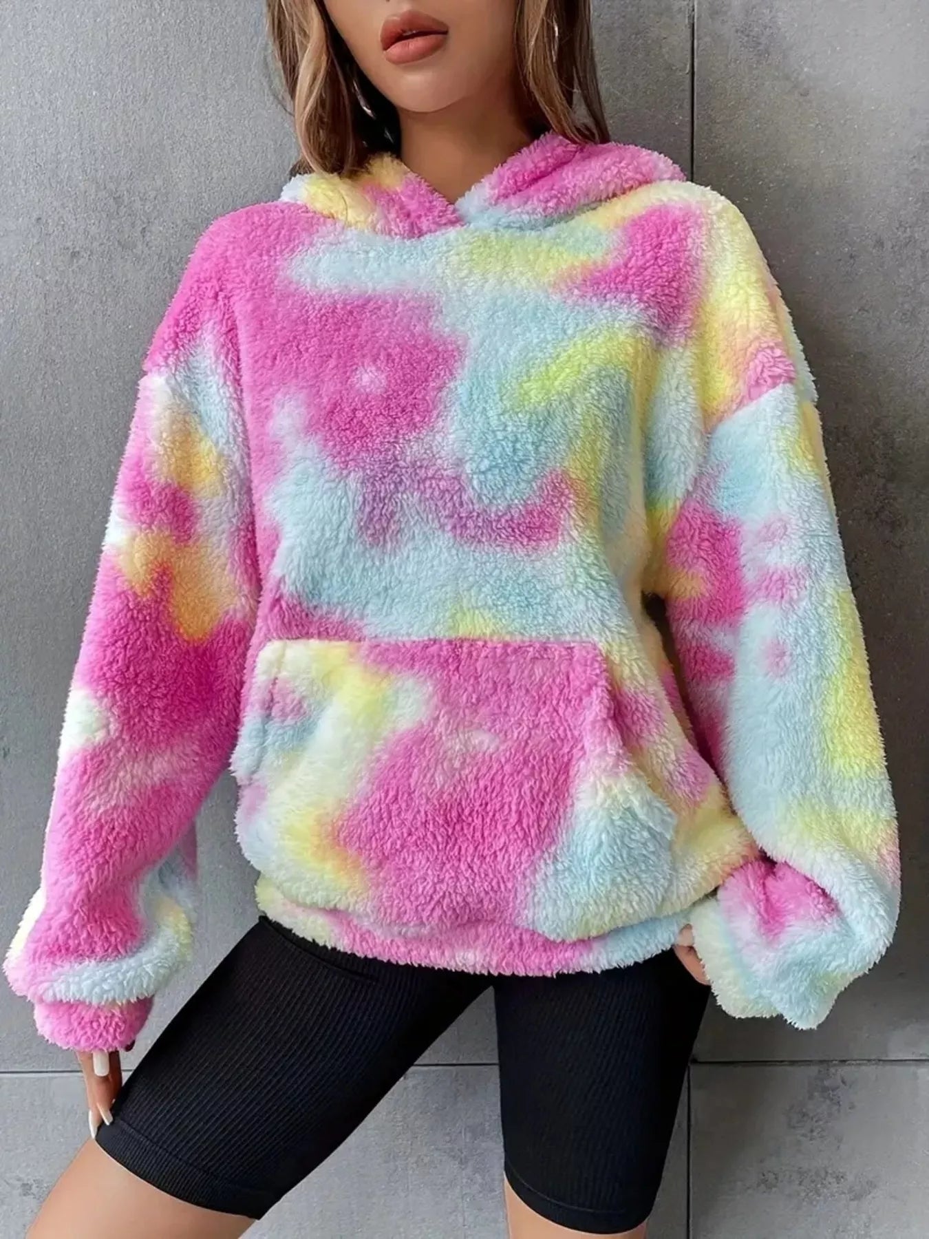 Model wearing a pastel tie-dye plush hoodie in pink, blue, and yellow with a kangaroo pocket and oversized cozy fit.