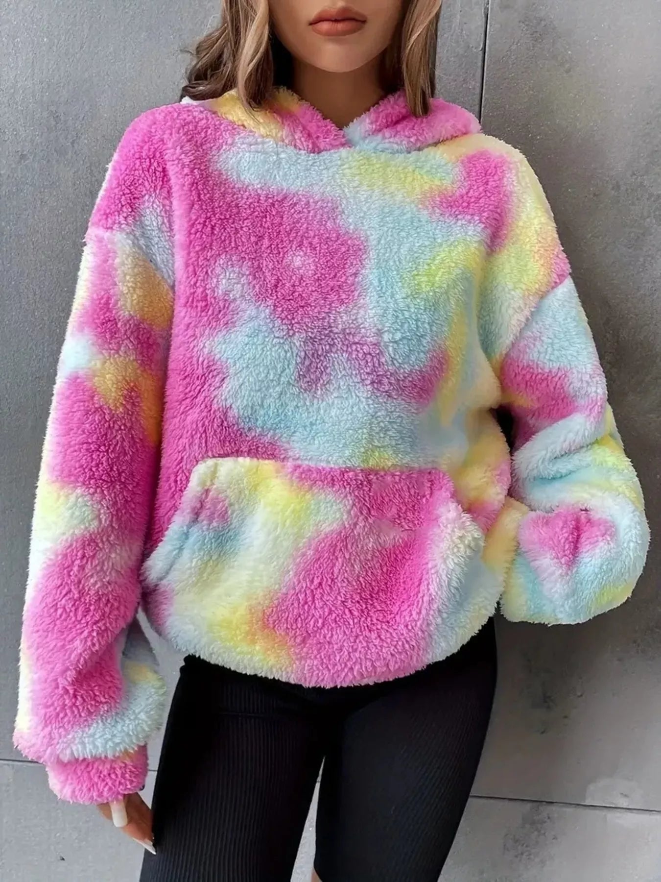 Model wearing a pastel tie-dye plush hoodie in pink, blue, and yellow with a kangaroo pocket and oversized cozy fit.