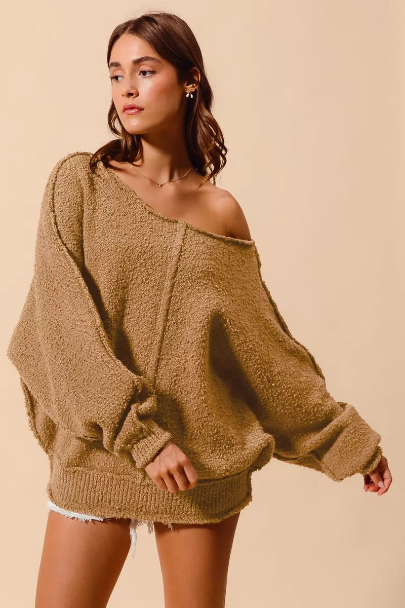Model wearing an oversized tan bouclé sweater with exposed seam detailing and slouchy off-the-shoulder fit, styled casually with shorts for a cozy and relaxed look.