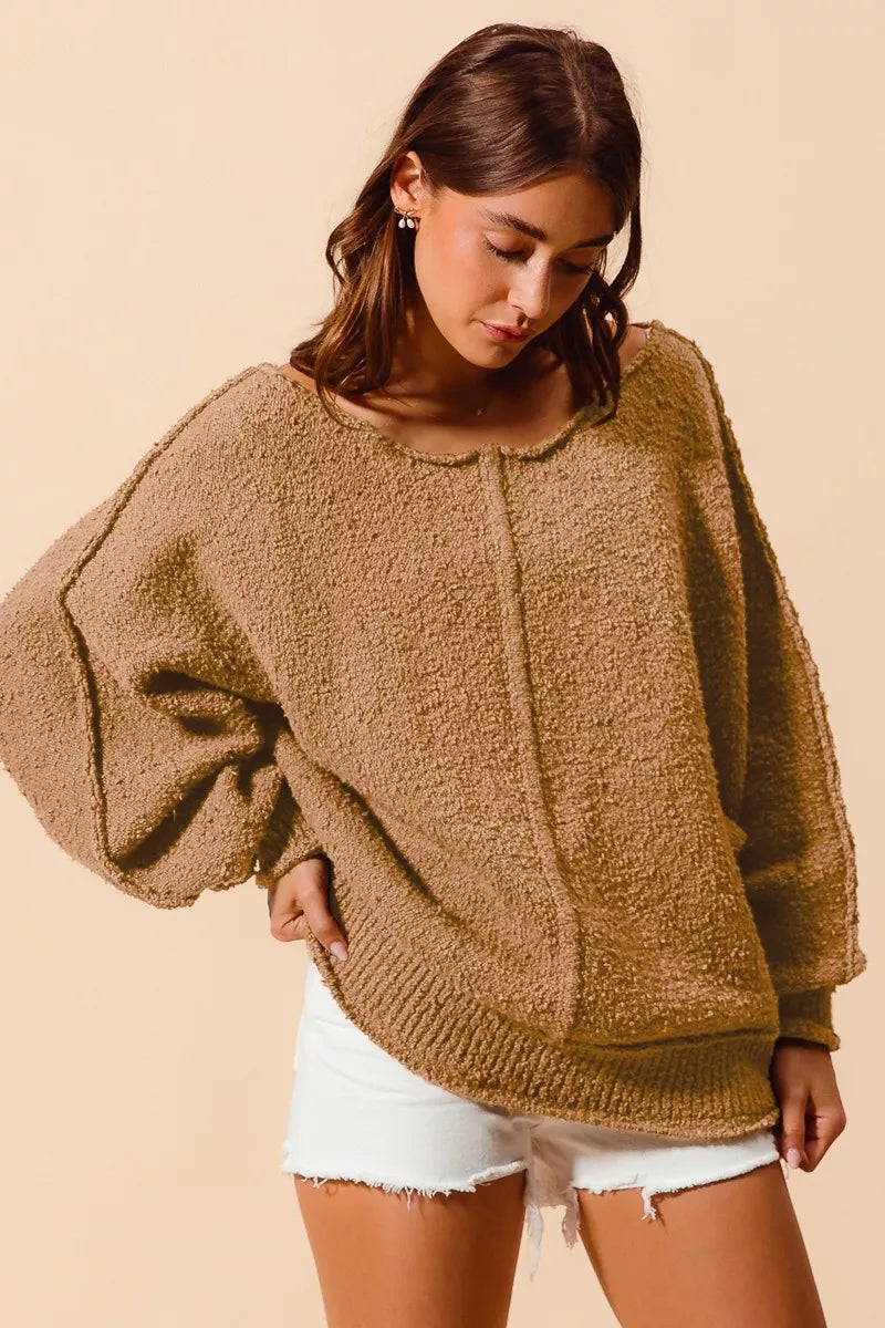 Model wearing an oversized tan bouclé sweater with exposed seam detailing and slouchy off-the-shoulder fit, styled casually with shorts for a cozy and relaxed look.