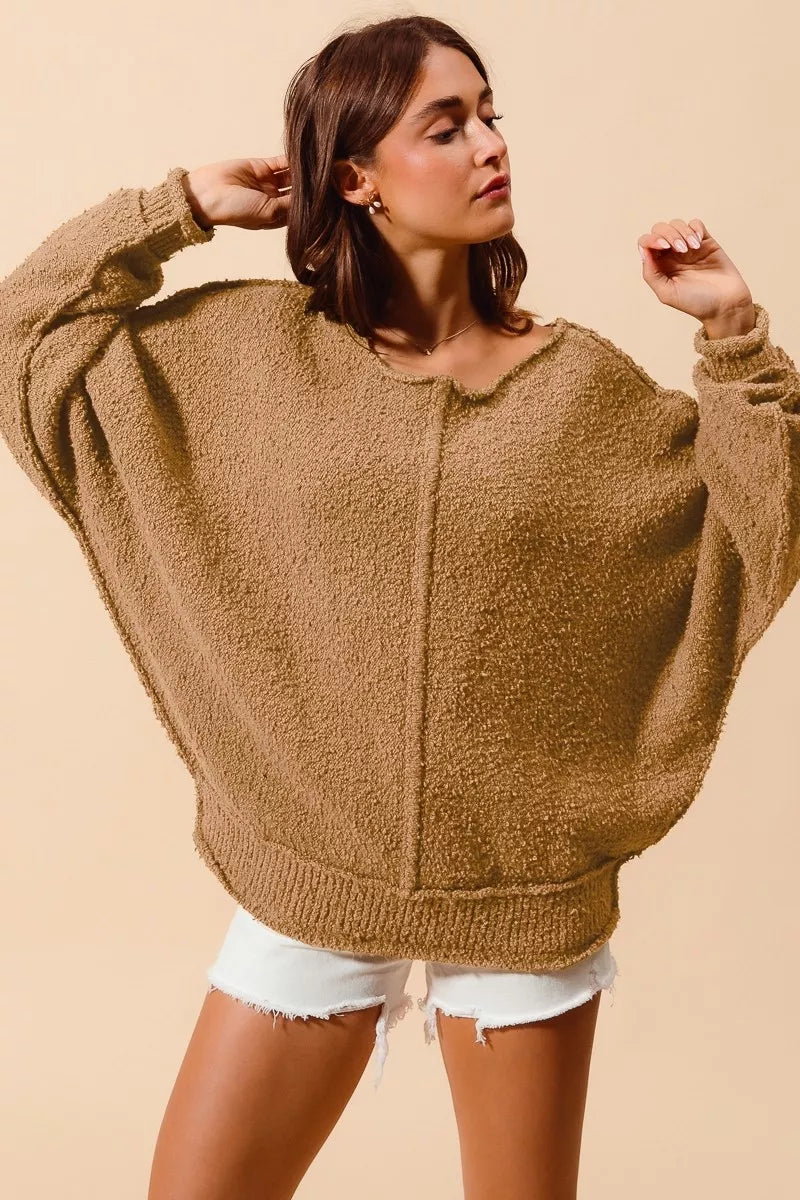 Model wearing an oversized tan bouclé sweater with exposed seam detailing and slouchy off-the-shoulder fit, styled casually with shorts for a cozy and relaxed look.