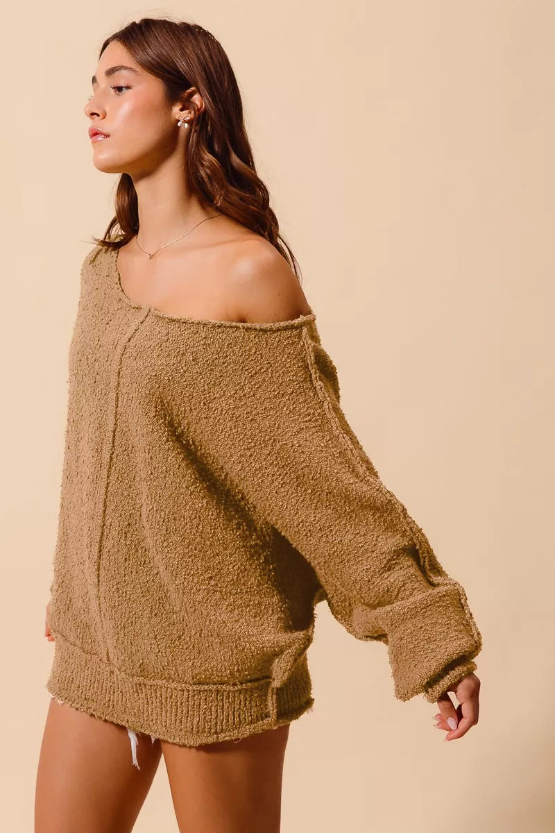 Model wearing an oversized tan bouclé sweater with exposed seam detailing and slouchy off-the-shoulder fit, styled casually with shorts for a cozy and relaxed look.