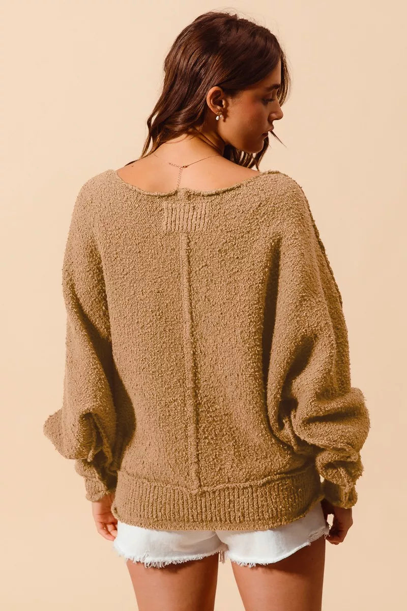 Model wearing an oversized tan bouclé sweater with exposed seam detailing and slouchy off-the-shoulder fit, styled casually with shorts for a cozy and relaxed look.
