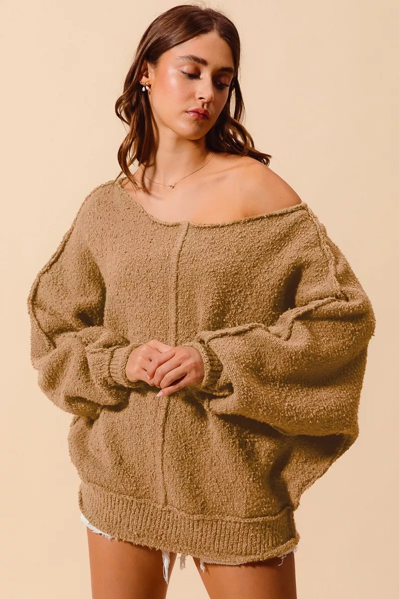 Model wearing an oversized tan bouclé sweater with exposed seam detailing and slouchy off-the-shoulder fit, styled casually with shorts for a cozy and relaxed look.
