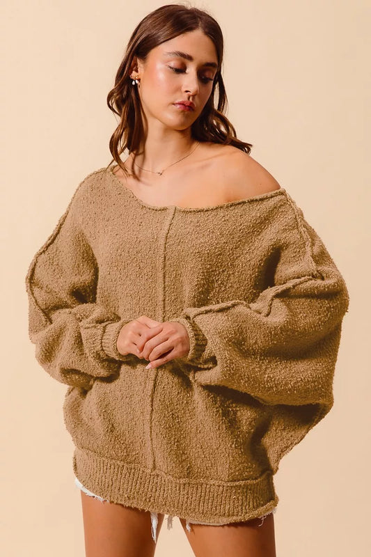 Model wearing an oversized tan bouclé sweater with exposed seam detailing and slouchy off-the-shoulder fit, styled casually with shorts for a cozy and relaxed look.
