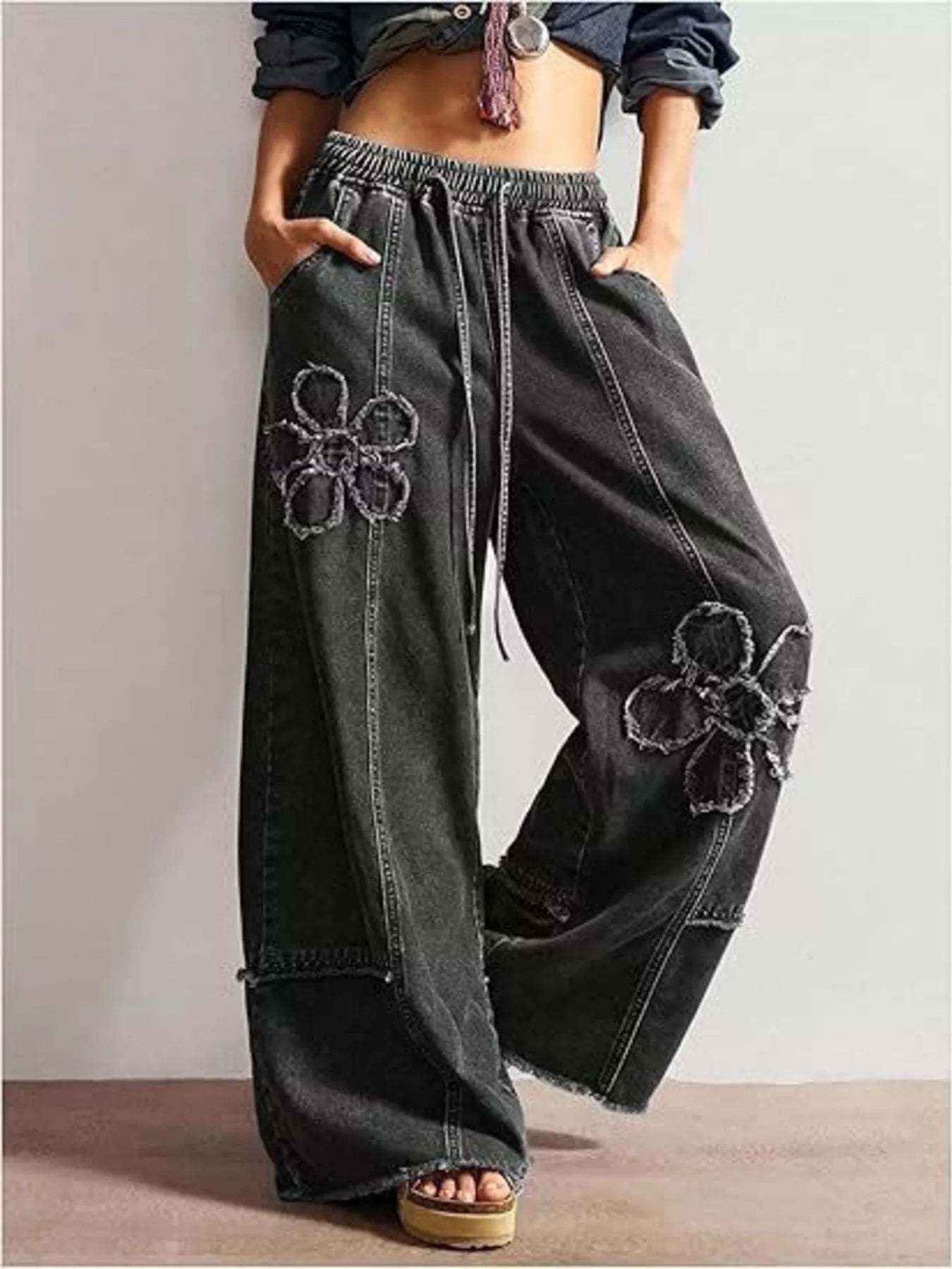 Wide-leg denim pants with drawstring waist, pockets, raw hem details, seam accents, and distressed floral appliqué designs.