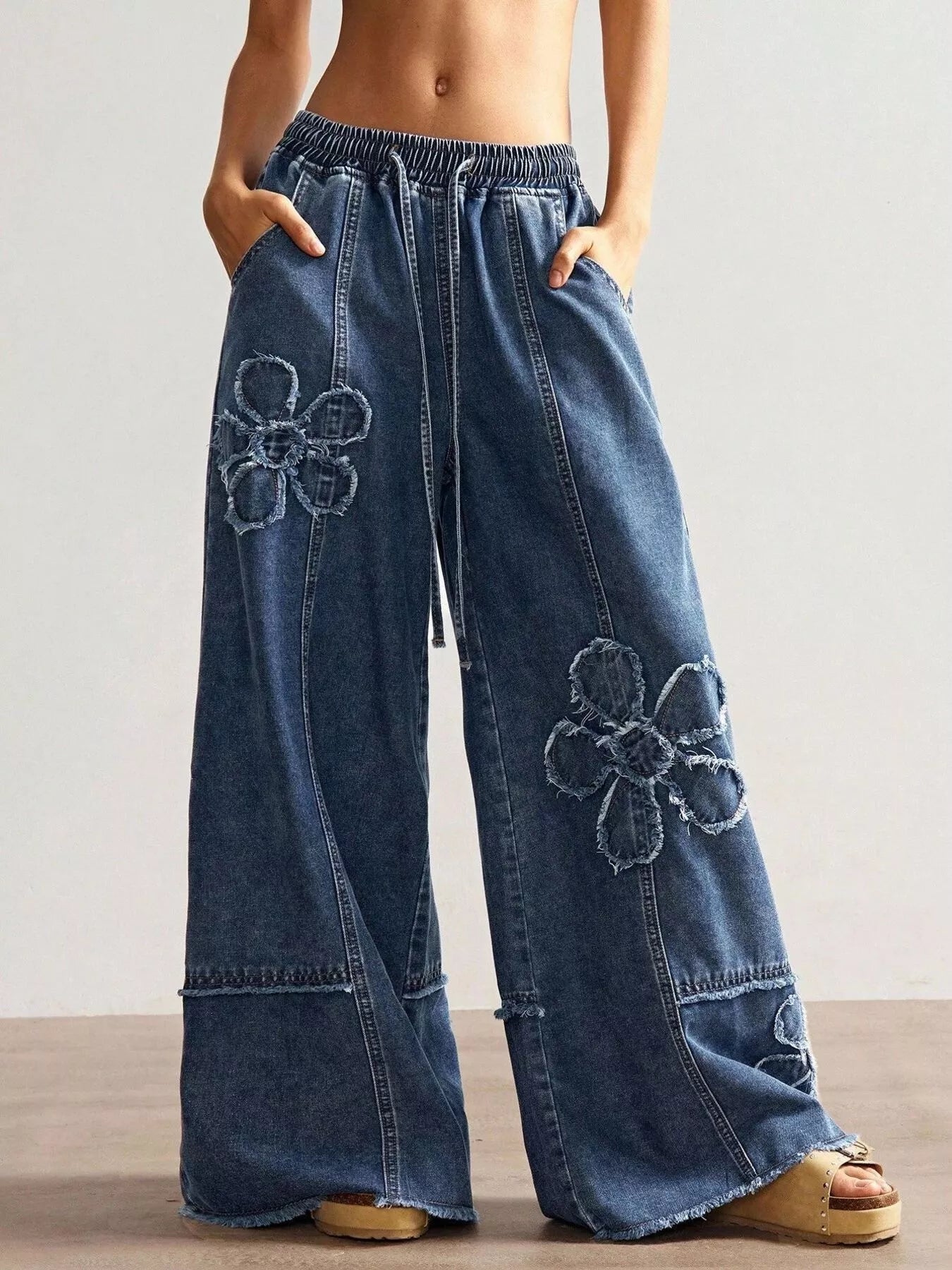 Wide-leg denim pants with drawstring waist, pockets, raw hem details, seam accents, and distressed floral appliqué designs.
