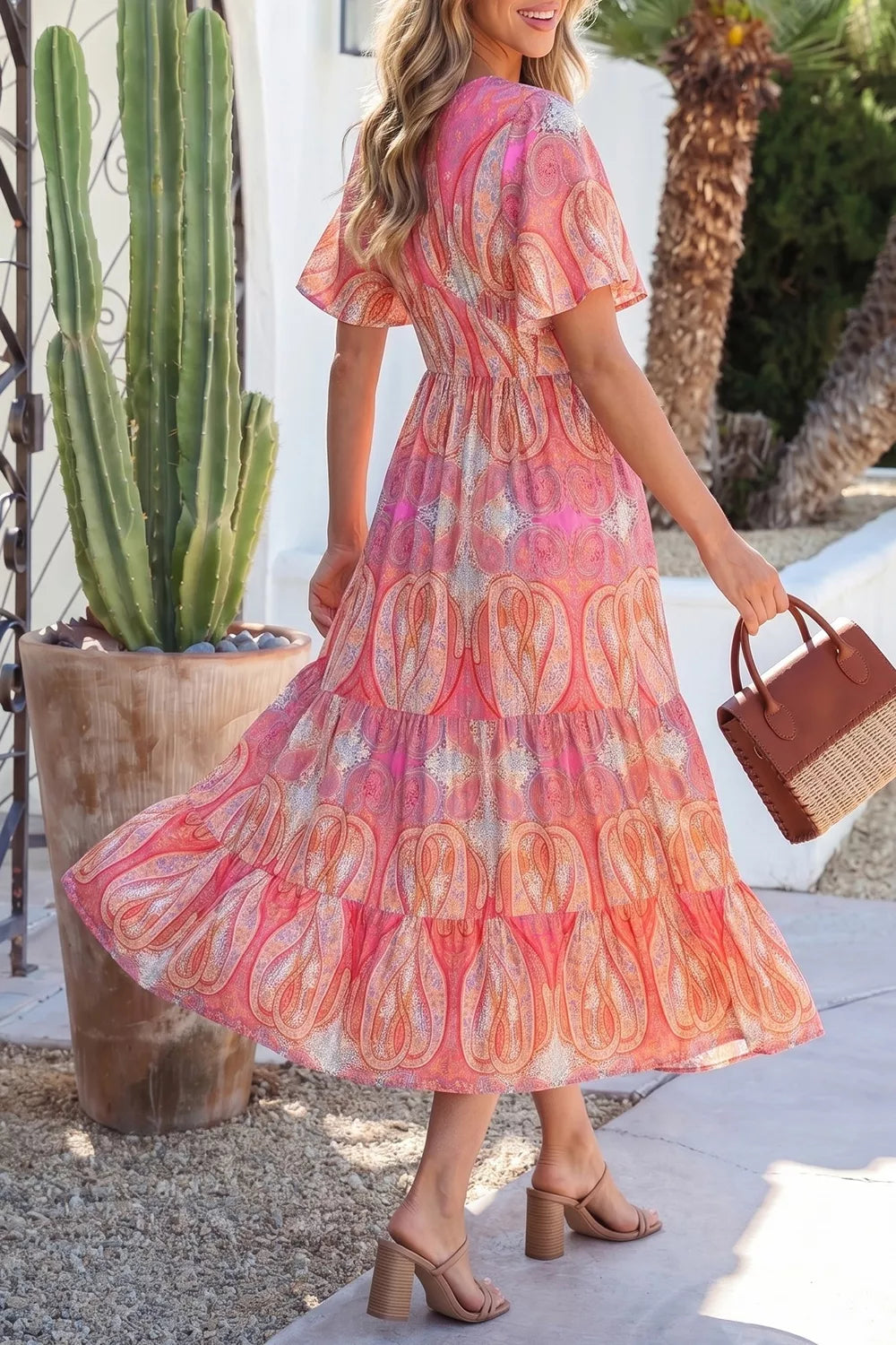 Pink paisley tiered midi dress with V-neckline, flutter sleeves, flowy silhouette, and slightly stretchy fabric styled with heels and a woven bag.