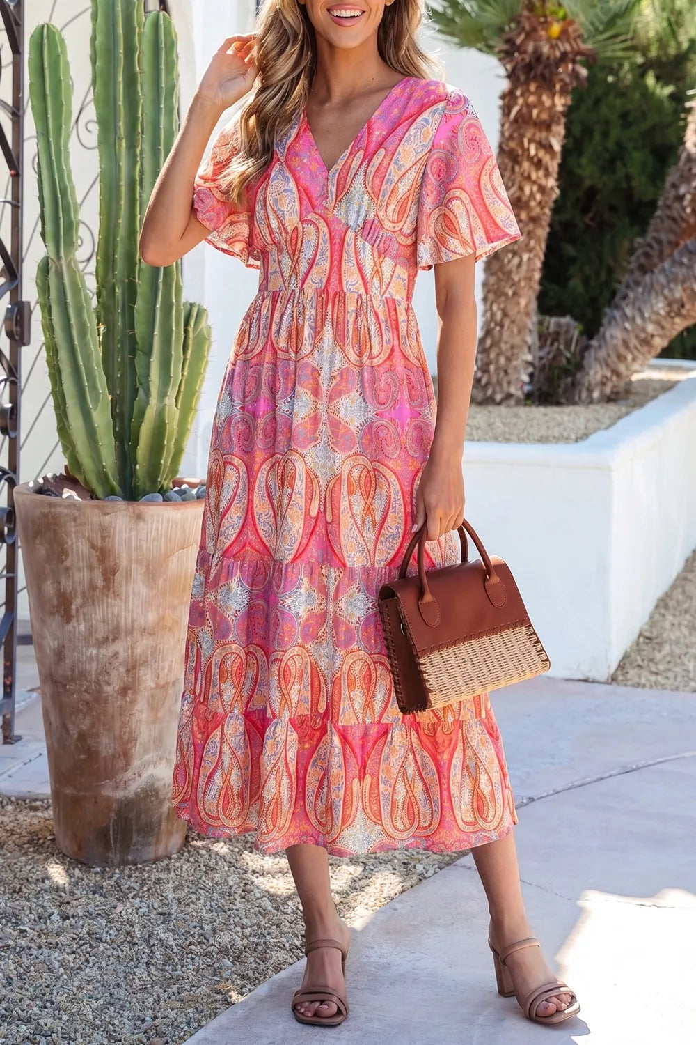 Pink paisley tiered midi dress with V-neckline, flutter sleeves, flowy silhouette, and slightly stretchy fabric styled with heels and a woven bag.