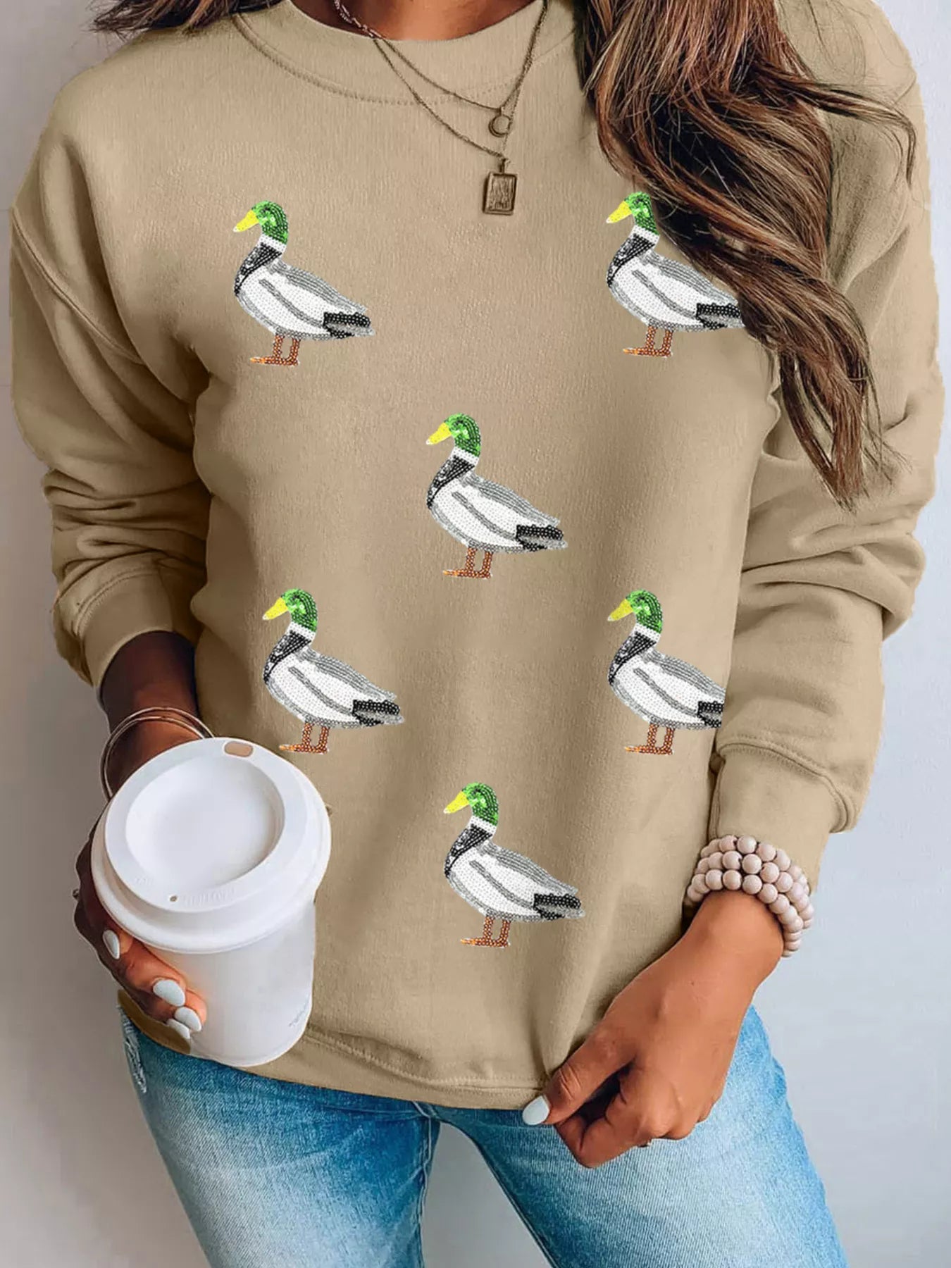 Woman wearing tan Duckie Sparkle Sweatshirt with sequin duck designs, fun quirky casual top for animal lovers.