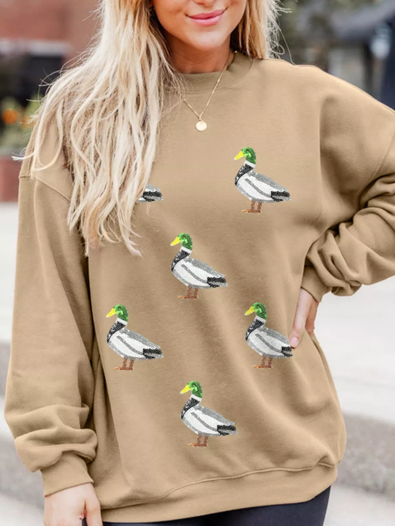 Woman wearing tan Duckie Sparkle Sweatshirt with sequin duck designs, fun quirky casual top for animal lovers.