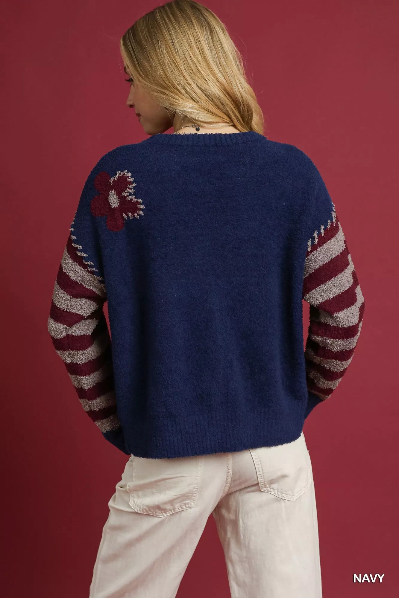 Model wearing a navy sweater with burgundy and gray striped sleeves, featuring a jacquard flower design and a relaxed fit, styled with light-colored pants.