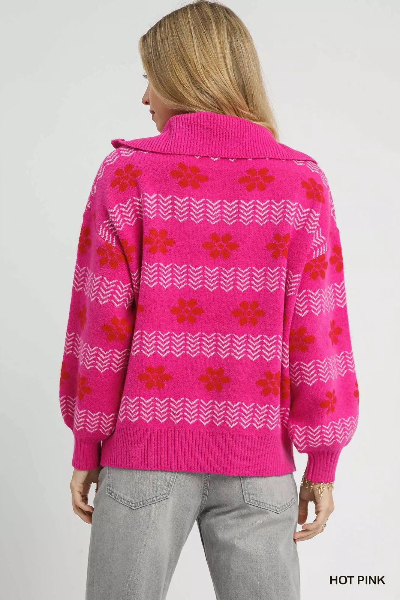 Woman wearing a hot pink floral and chevron jacquard quarter-zip sweater with ribbed cuffs and hem, paired with light grey jeans.