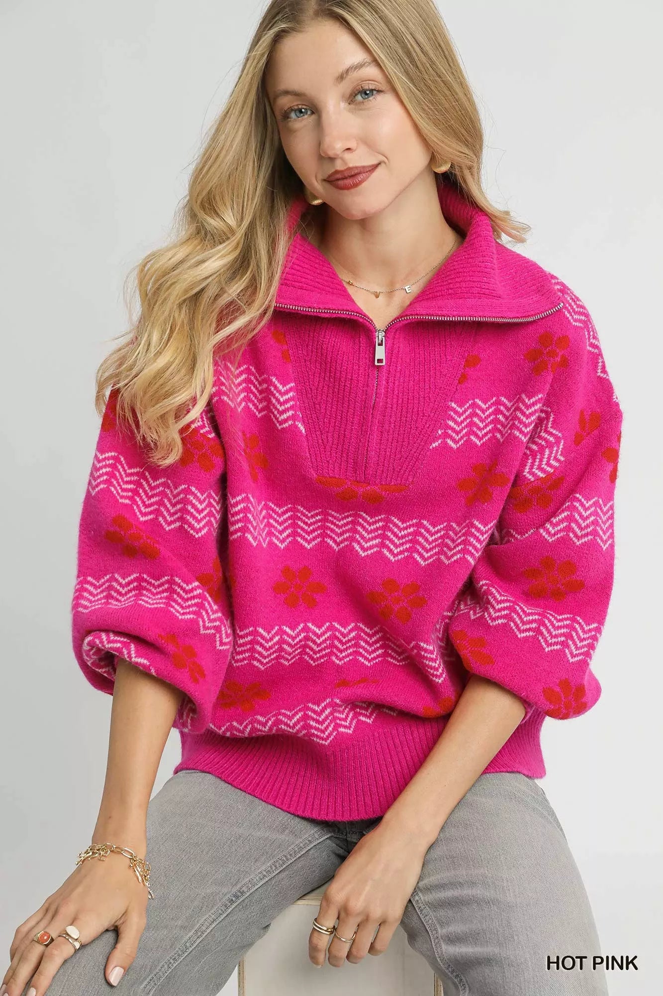 Woman wearing a hot pink floral and chevron jacquard quarter-zip sweater with ribbed cuffs and hem, paired with light grey jeans.