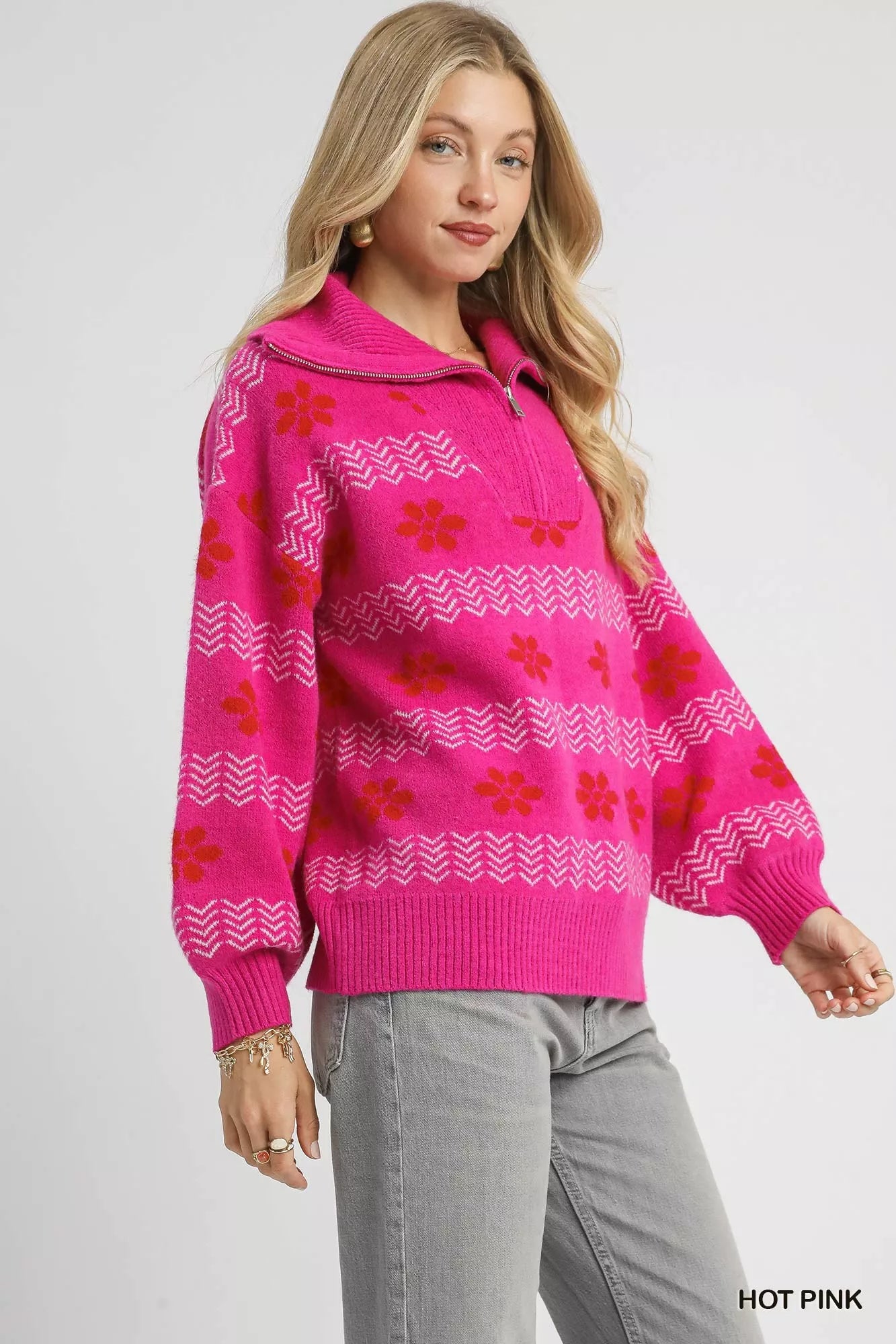 Woman wearing a hot pink floral and chevron jacquard quarter-zip sweater with ribbed cuffs and hem, paired with light grey jeans.