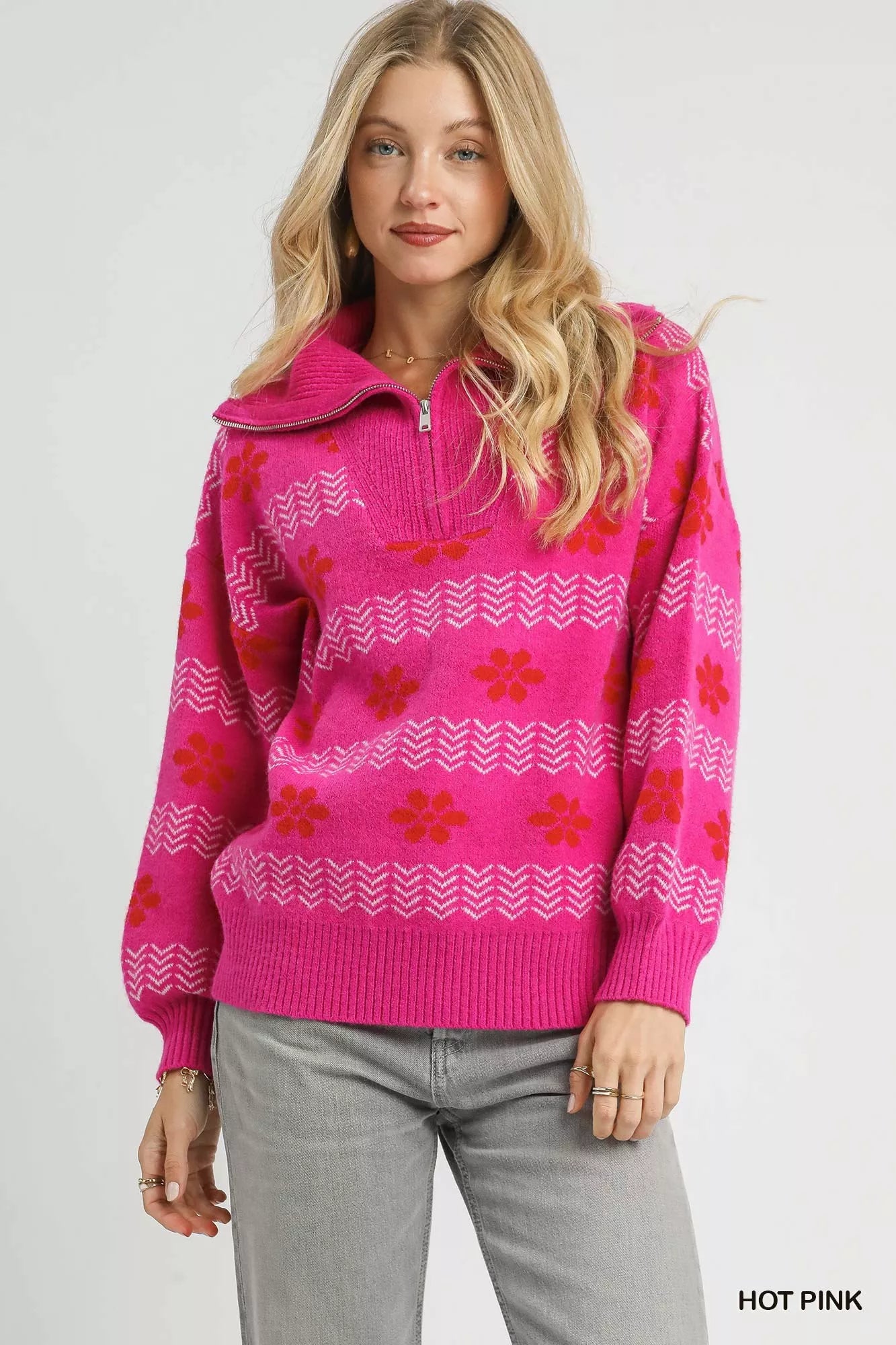 Woman wearing a hot pink floral and chevron jacquard quarter-zip sweater with ribbed cuffs and hem, paired with light grey jeans.