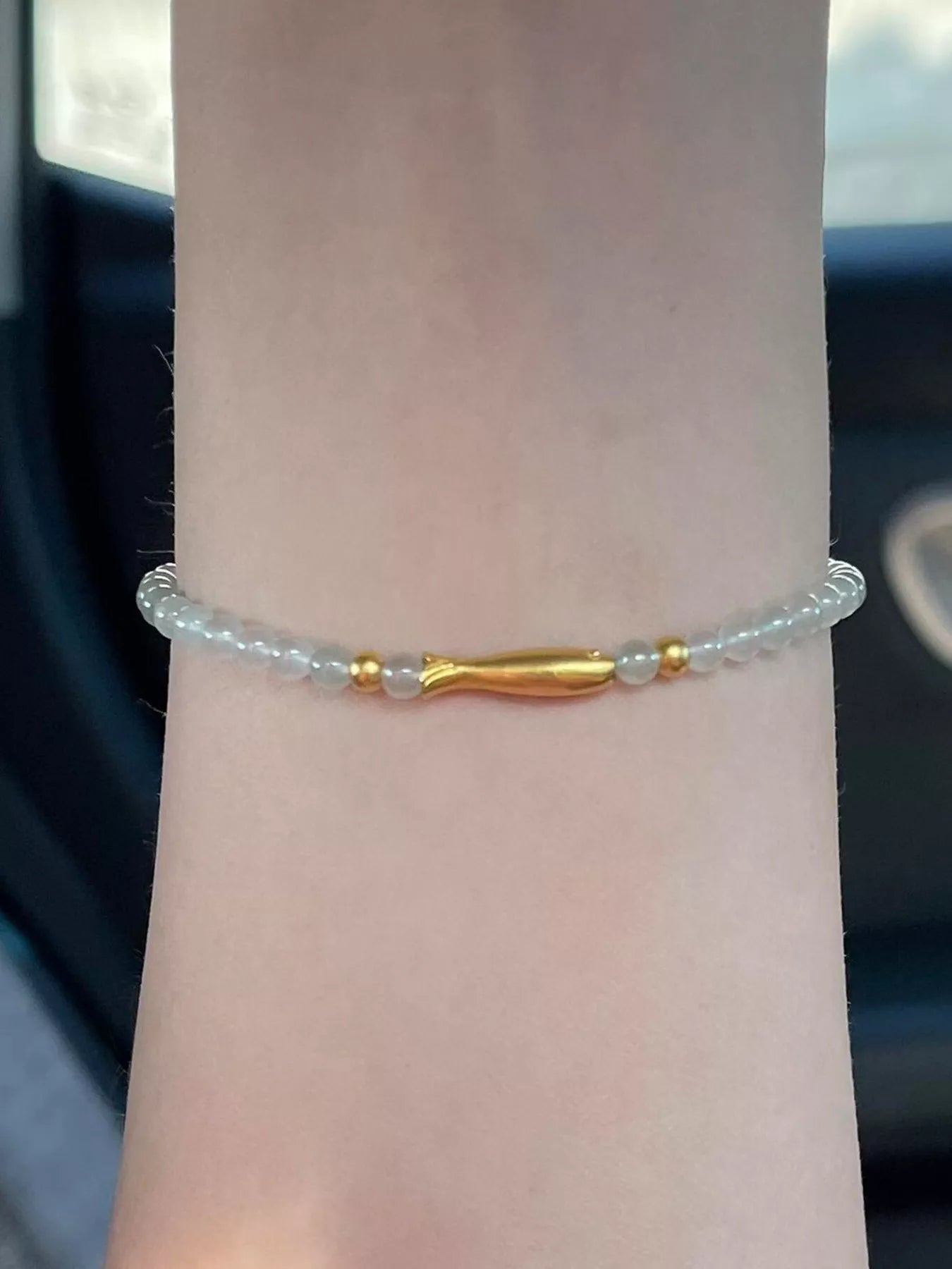 Dainty beaded bracelet with pale glass beads, 18K gold-plated accents, and a gold fish charm worn on the wrist.