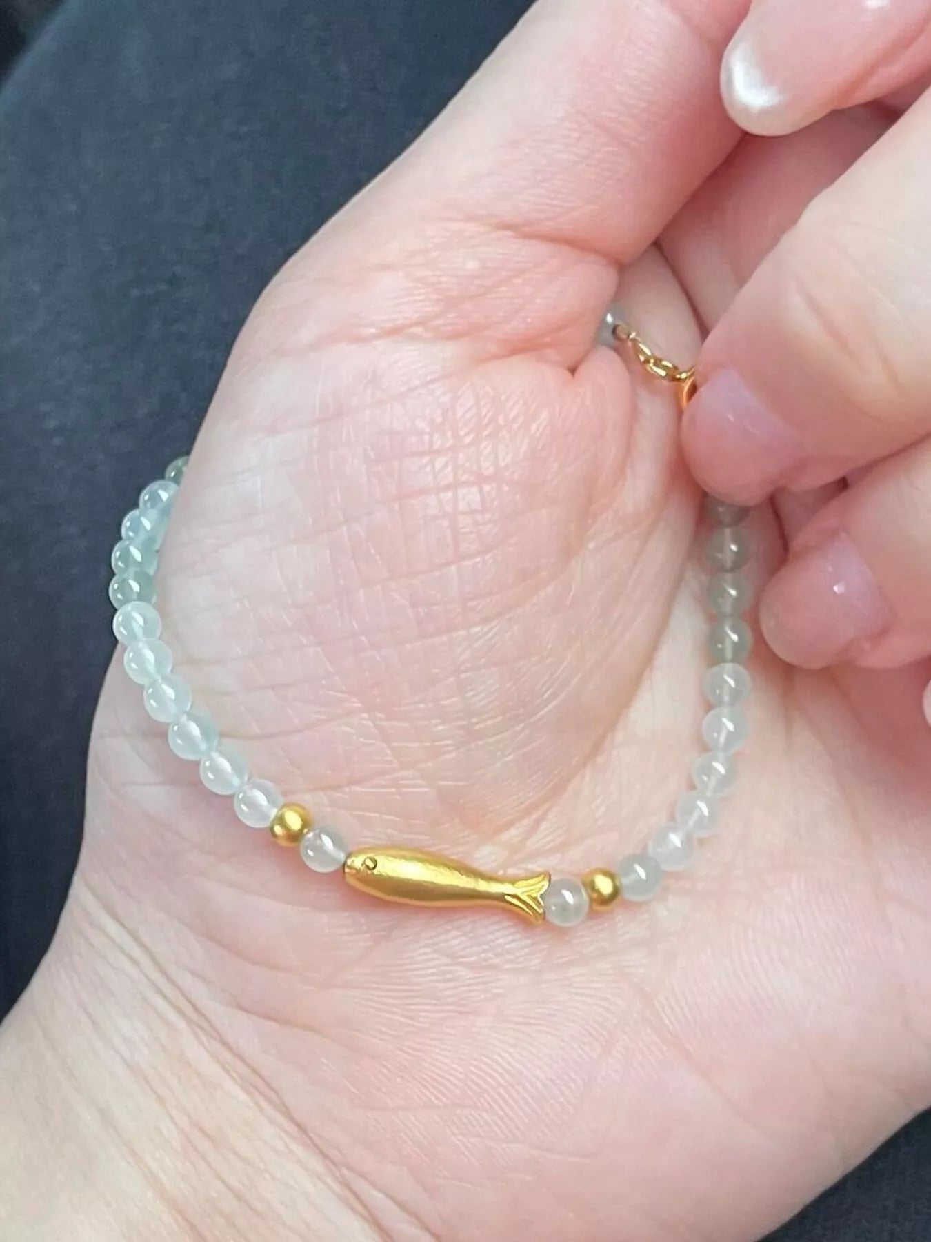 Dainty beaded bracelet with pale glass beads, 18K gold-plated accents, and a gold fish charm worn on the wrist.