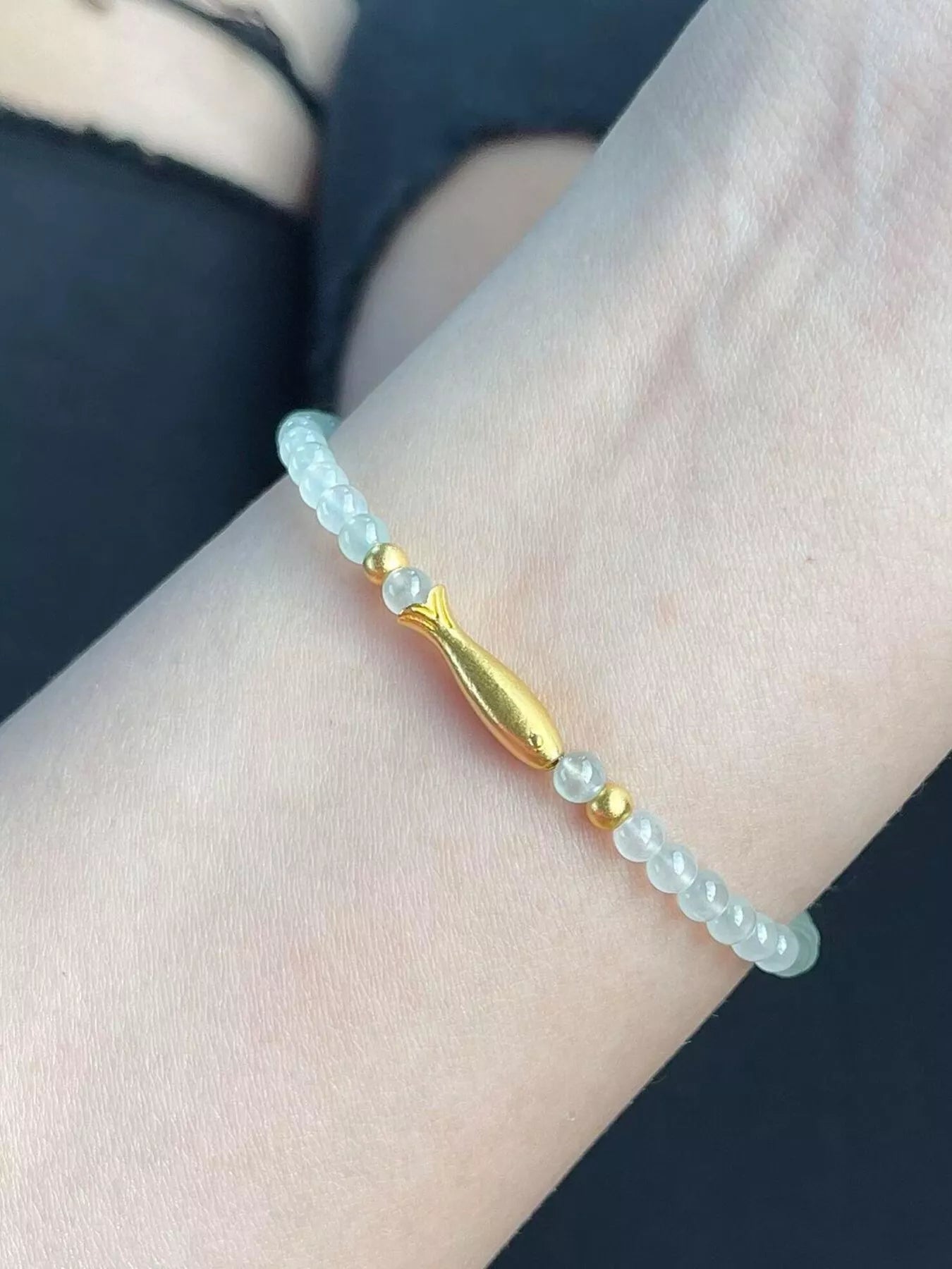 Dainty beaded bracelet with pale glass beads, 18K gold-plated accents, and a gold fish charm worn on the wrist.
