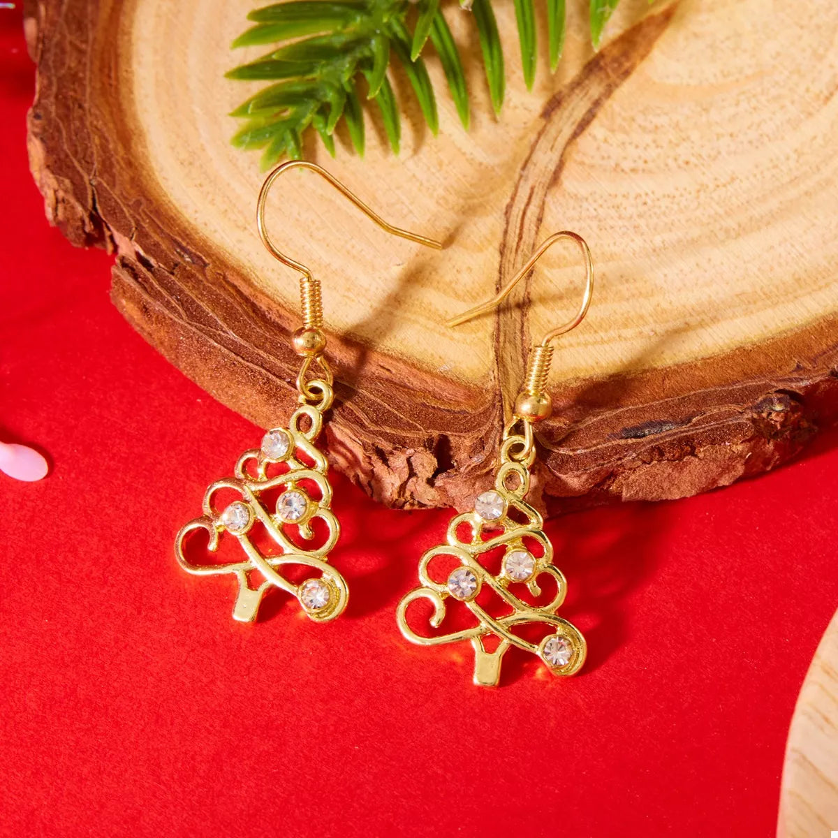 Gold Christmas tree-shaped earrings with rhinestone accents hanging on a gold display bar against a red background.
