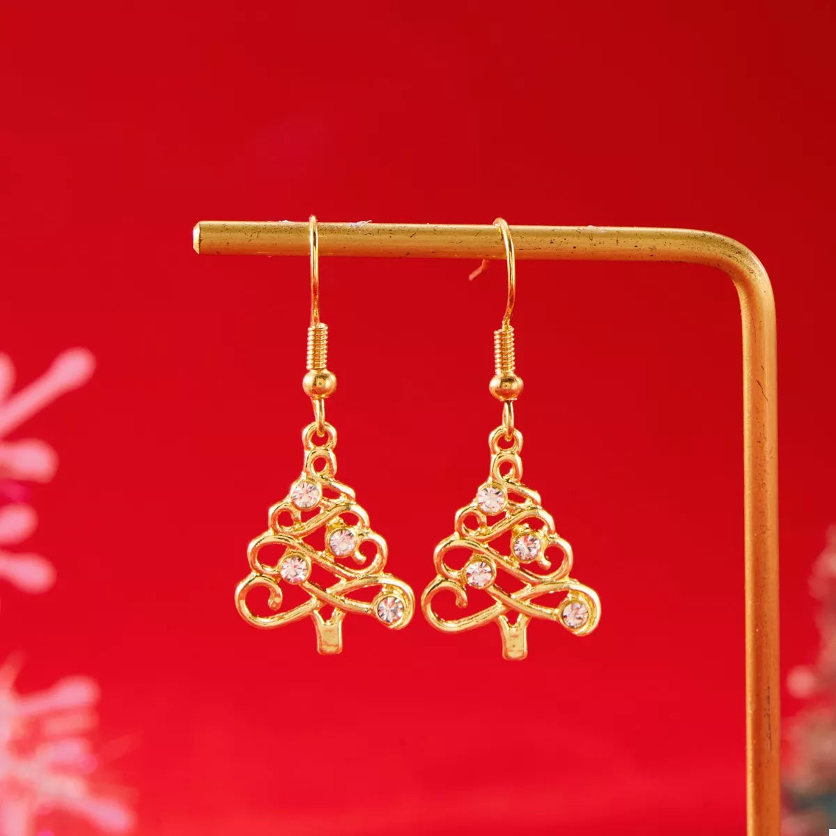 Gold Christmas tree-shaped earrings with rhinestone accents hanging on a gold display bar against a red background.
