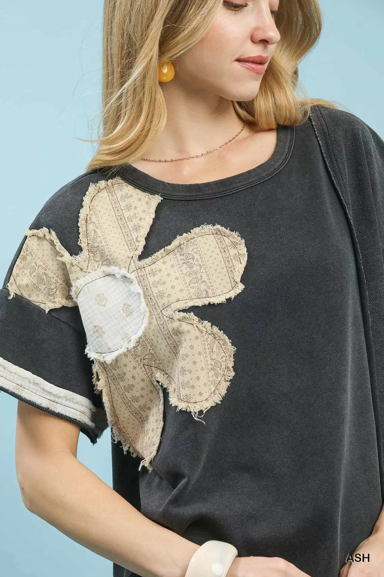 Woman wearing an ash-colored French terry top with floral patch, contrast panels, and exposed seams