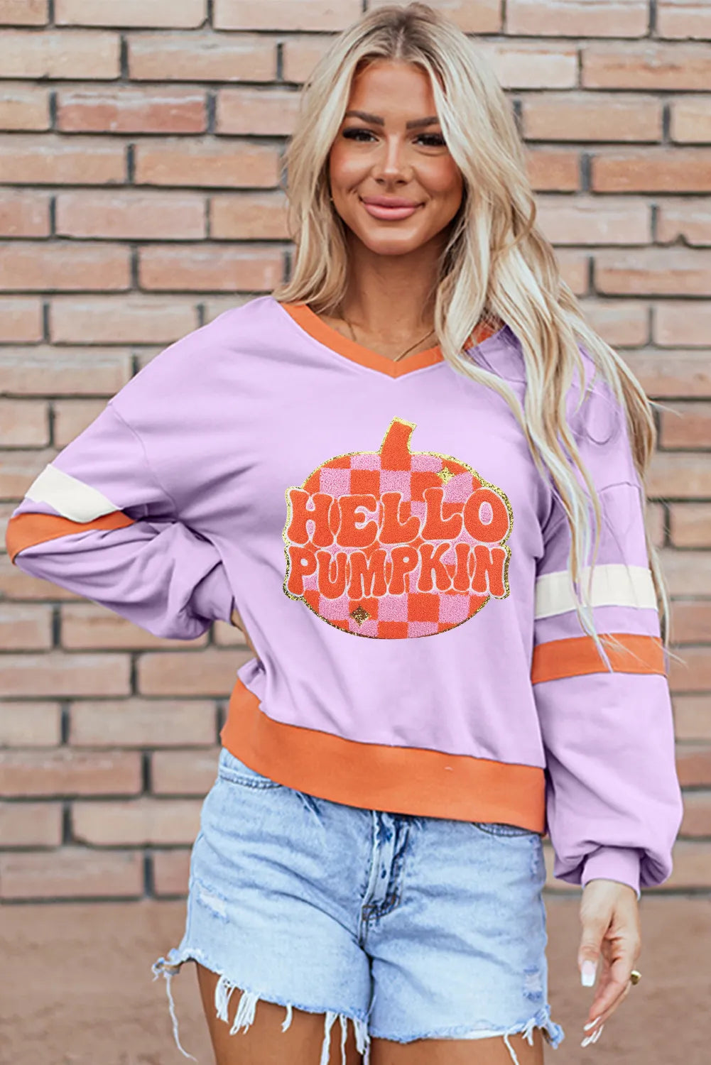 Woman wearing a lilac sweatshirt with a glittery orange checkered pumpkin and the phrase “Hello Pumpkin,” styled with denim shorts — cozy fall sweatshirt from A Whimsical Affair.