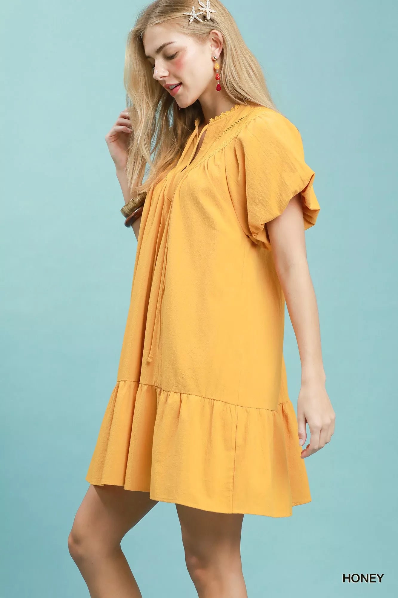 Honey yellow cotton mini dress with split V-neckline, tassel tie detail, flutter sleeves, ruffled hem, and side pockets.