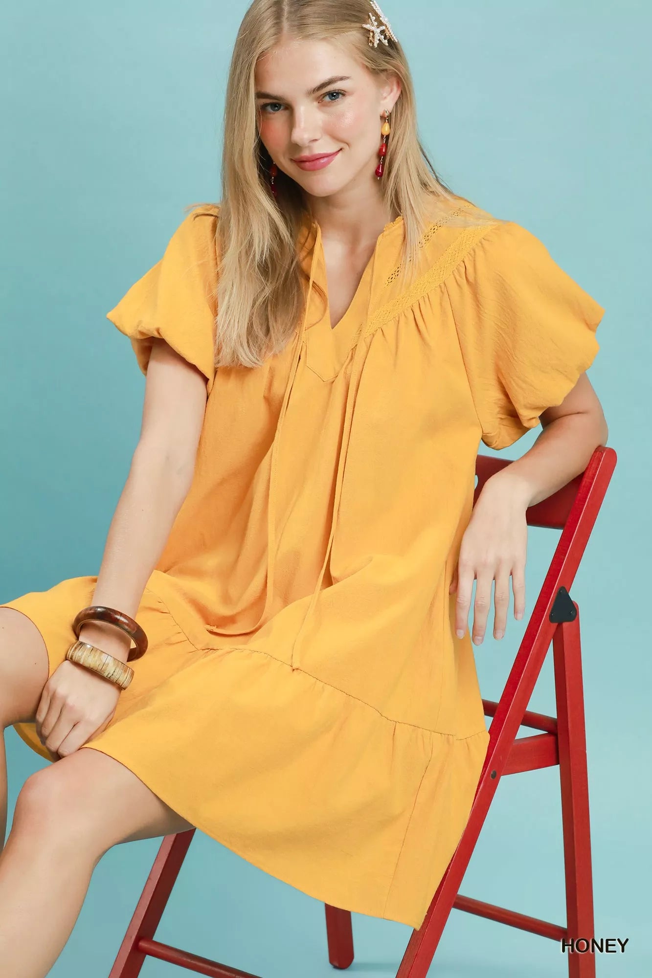 Honey yellow cotton mini dress with split V-neckline, tassel tie detail, flutter sleeves, ruffled hem, and side pockets.
