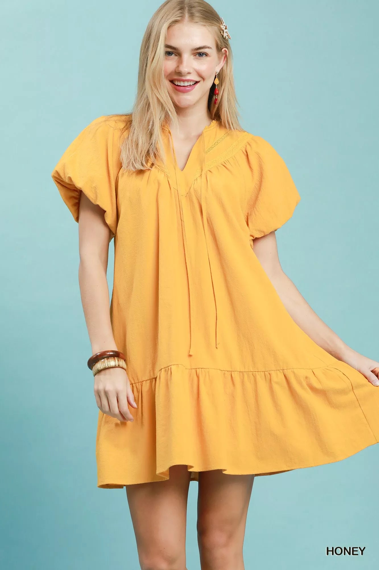 Honey yellow cotton mini dress with split V-neckline, tassel tie detail, flutter sleeves, ruffled hem, and side pockets.
