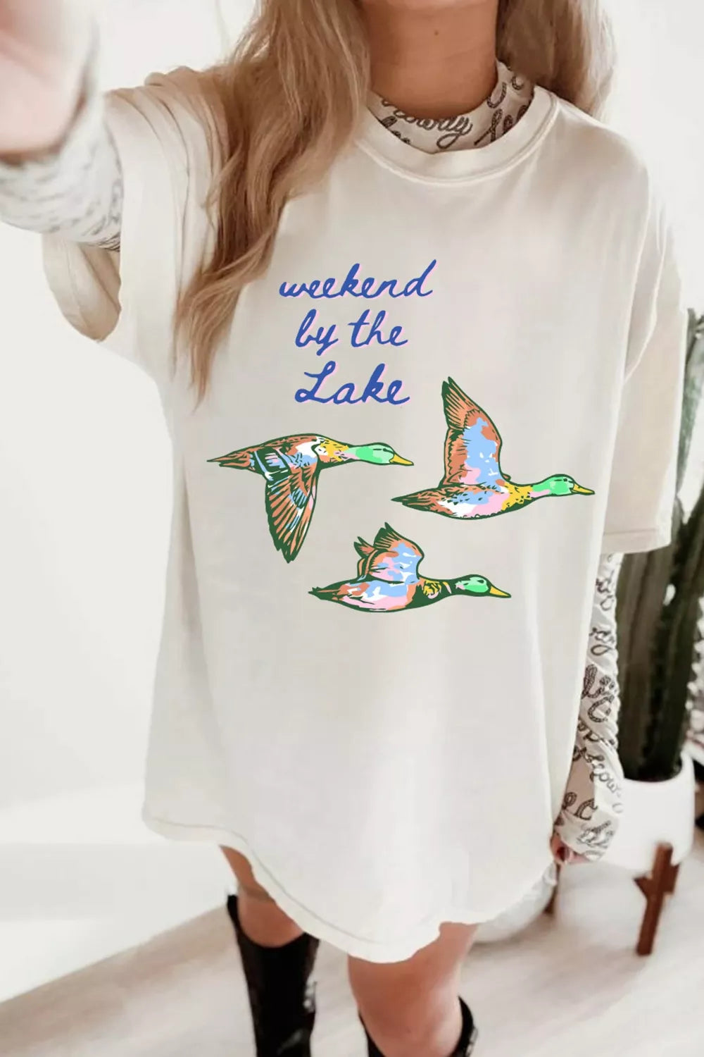 White graphic tee with “weekend by the lake” text and colorful flying duck design on the front.