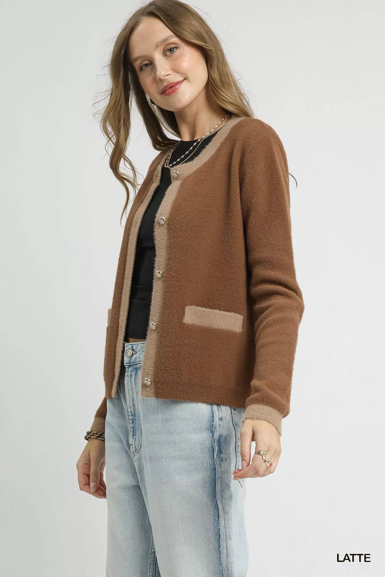 Model wearing a latte-colored two-tone cardigan with metallic buttons and contrast trim pockets, styled with a black top and light denim jeans.