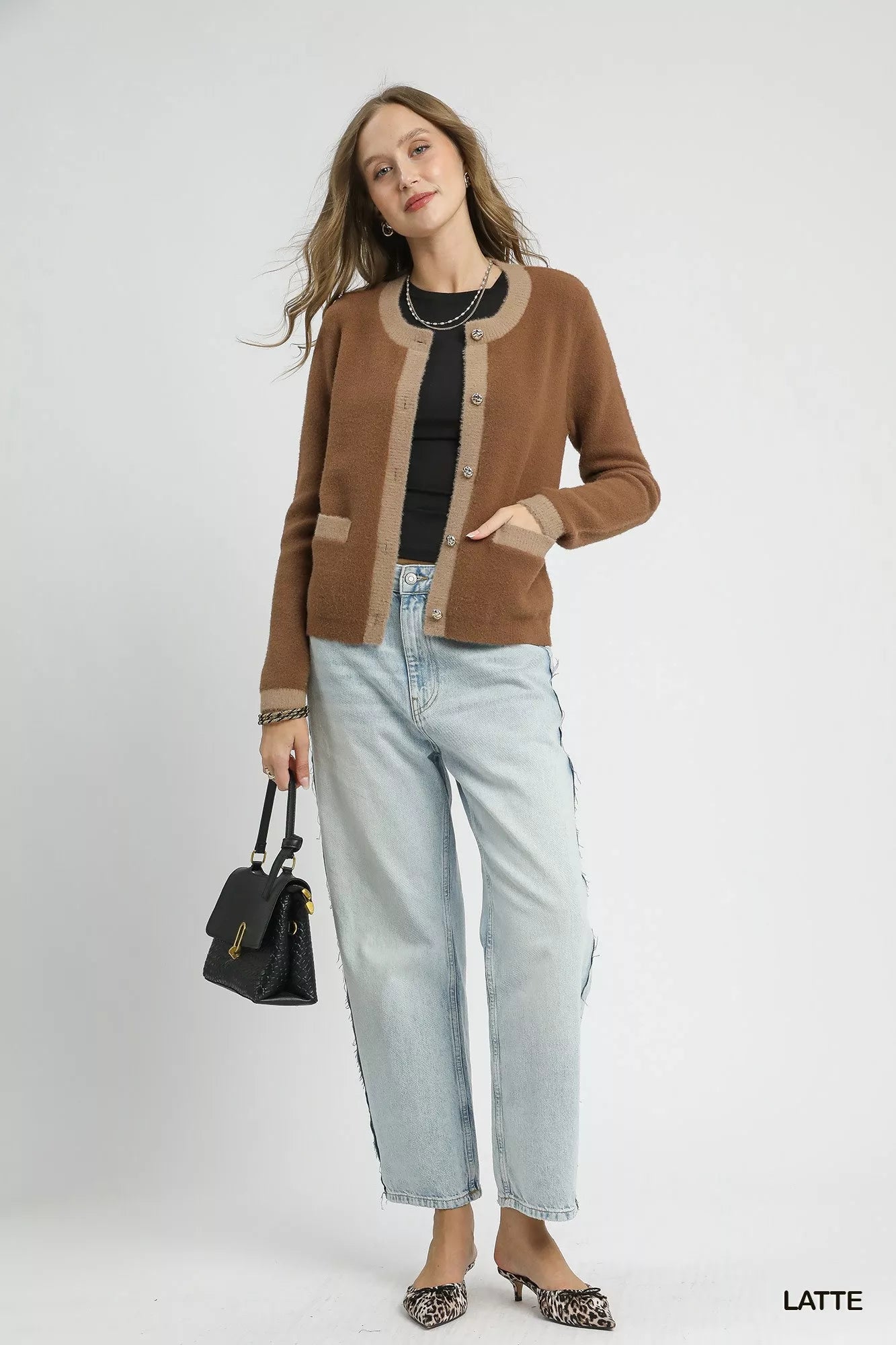 Model wearing a latte-colored two-tone cardigan with metallic buttons and contrast trim pockets, styled with a black top and light denim jeans.
