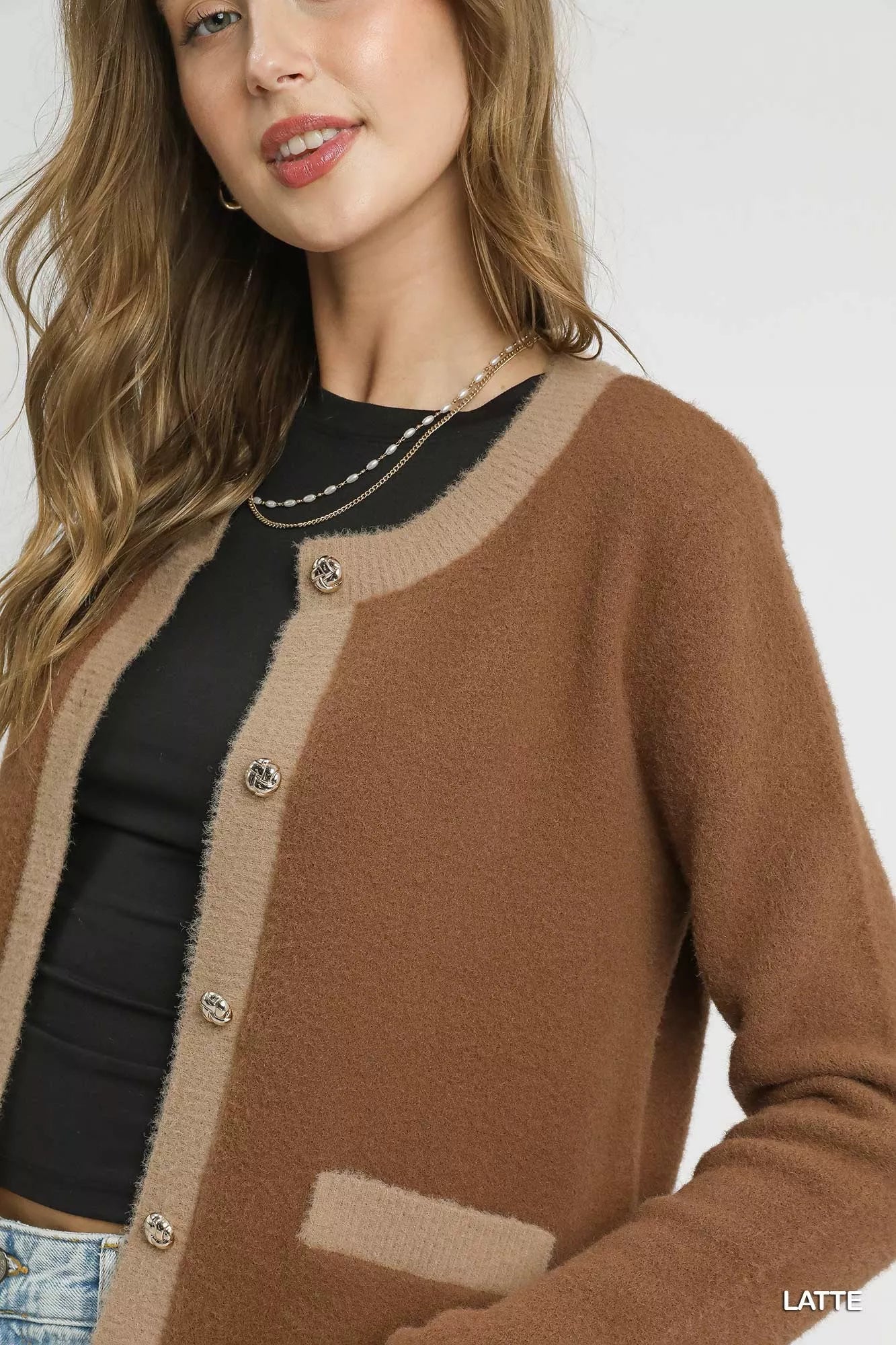 Model wearing a latte-colored two-tone cardigan with metallic buttons and contrast trim pockets, styled with a black top and light denim jeans.