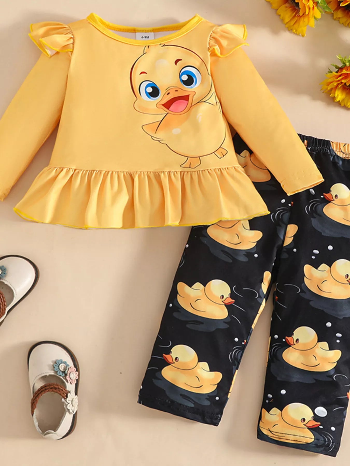 Two-piece baby outfit featuring a yellow long-sleeve ruffled top with a cute duck print and black pants with yellow duck patterns, displayed with floral sandals.