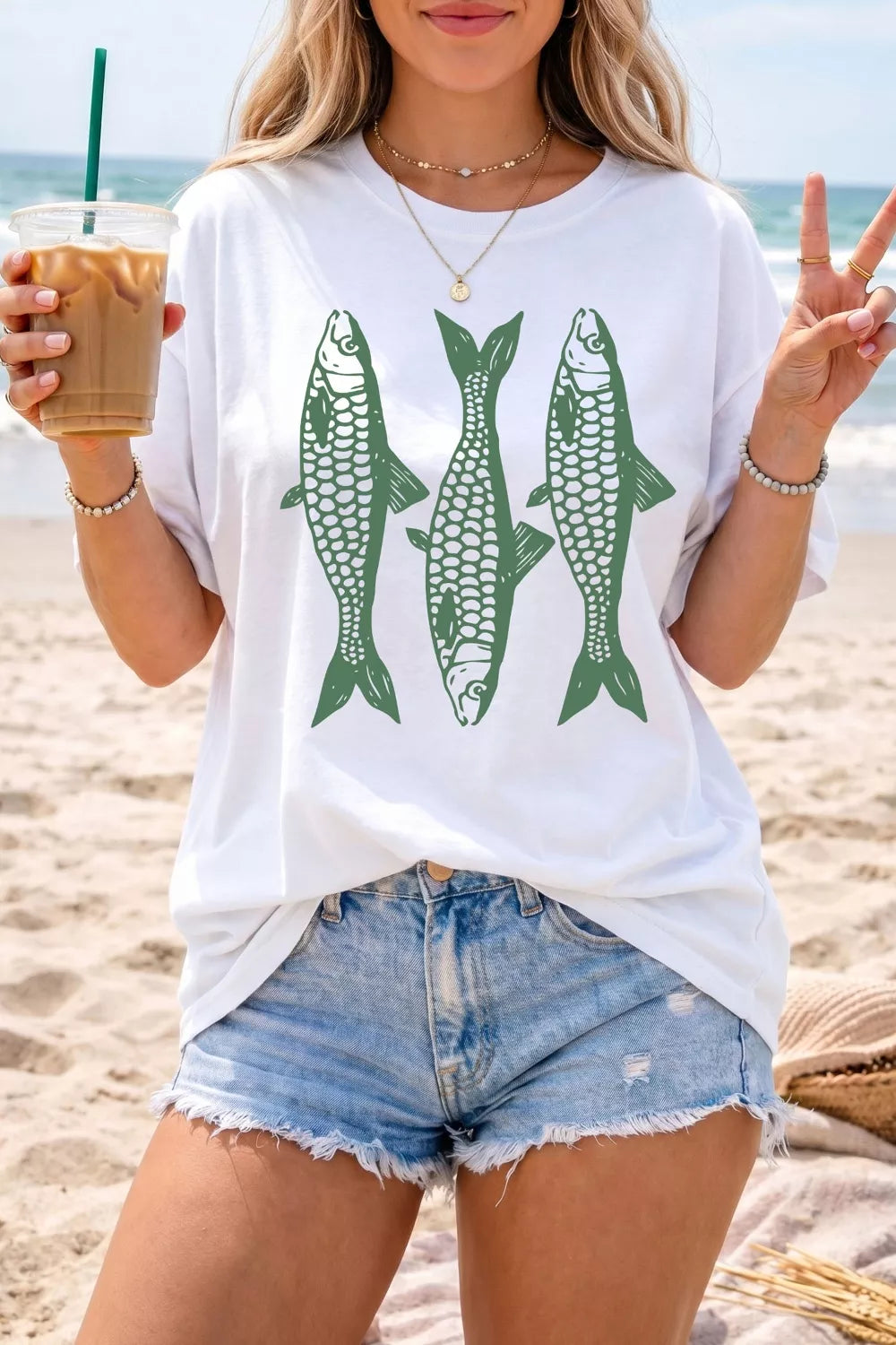 White short sleeve graphic tee with three green fish illustrations on the front, styled with denim jeans and a green bag.