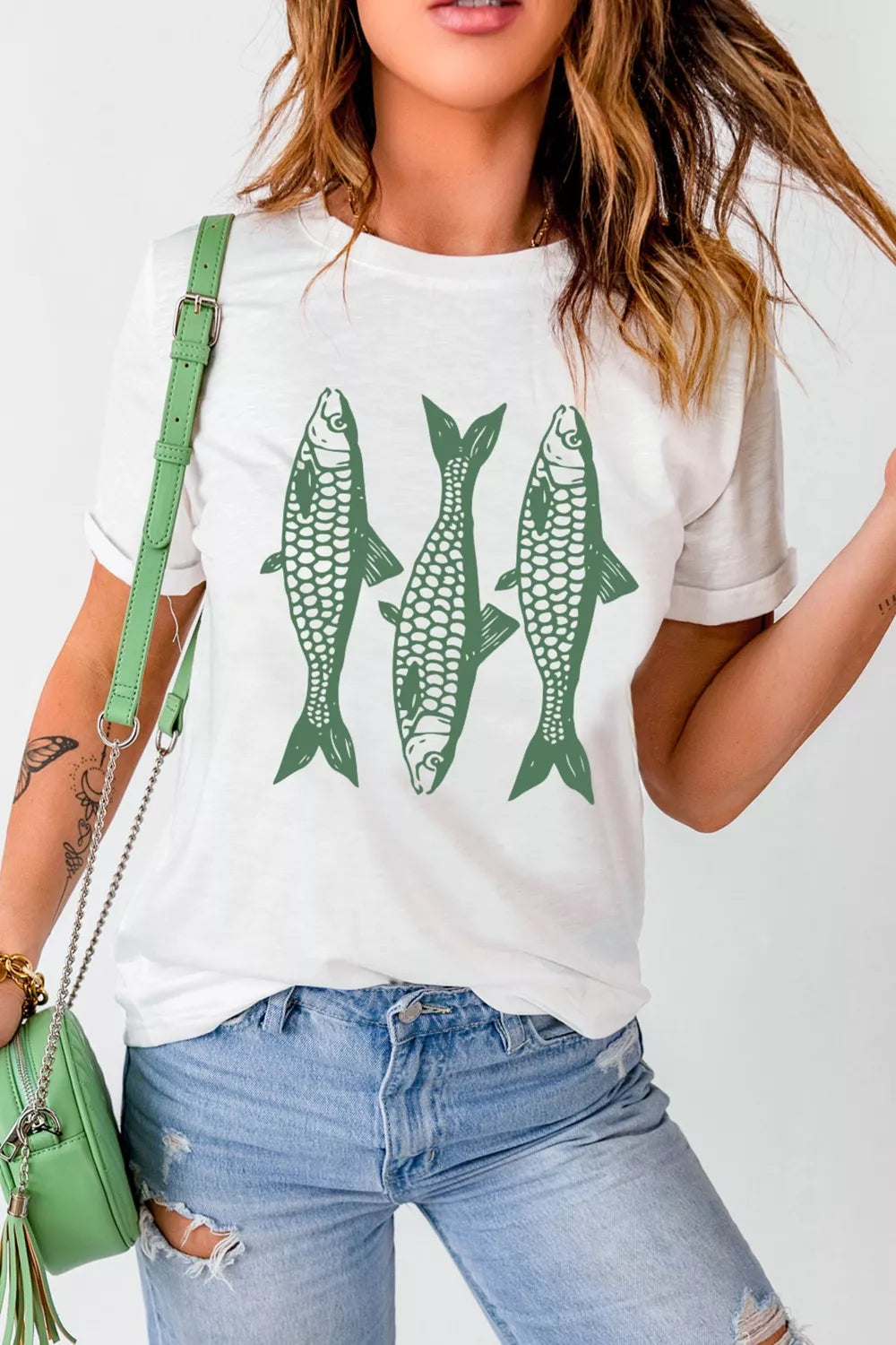White short sleeve graphic tee with three green fish illustrations on the front, styled with denim jeans and a green bag.