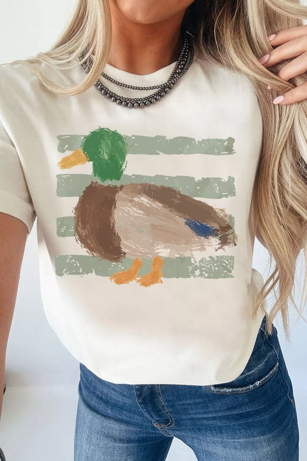 White short sleeve graphic tee with a painterly mallard duck design over muted green stripes, styled with camo shorts.