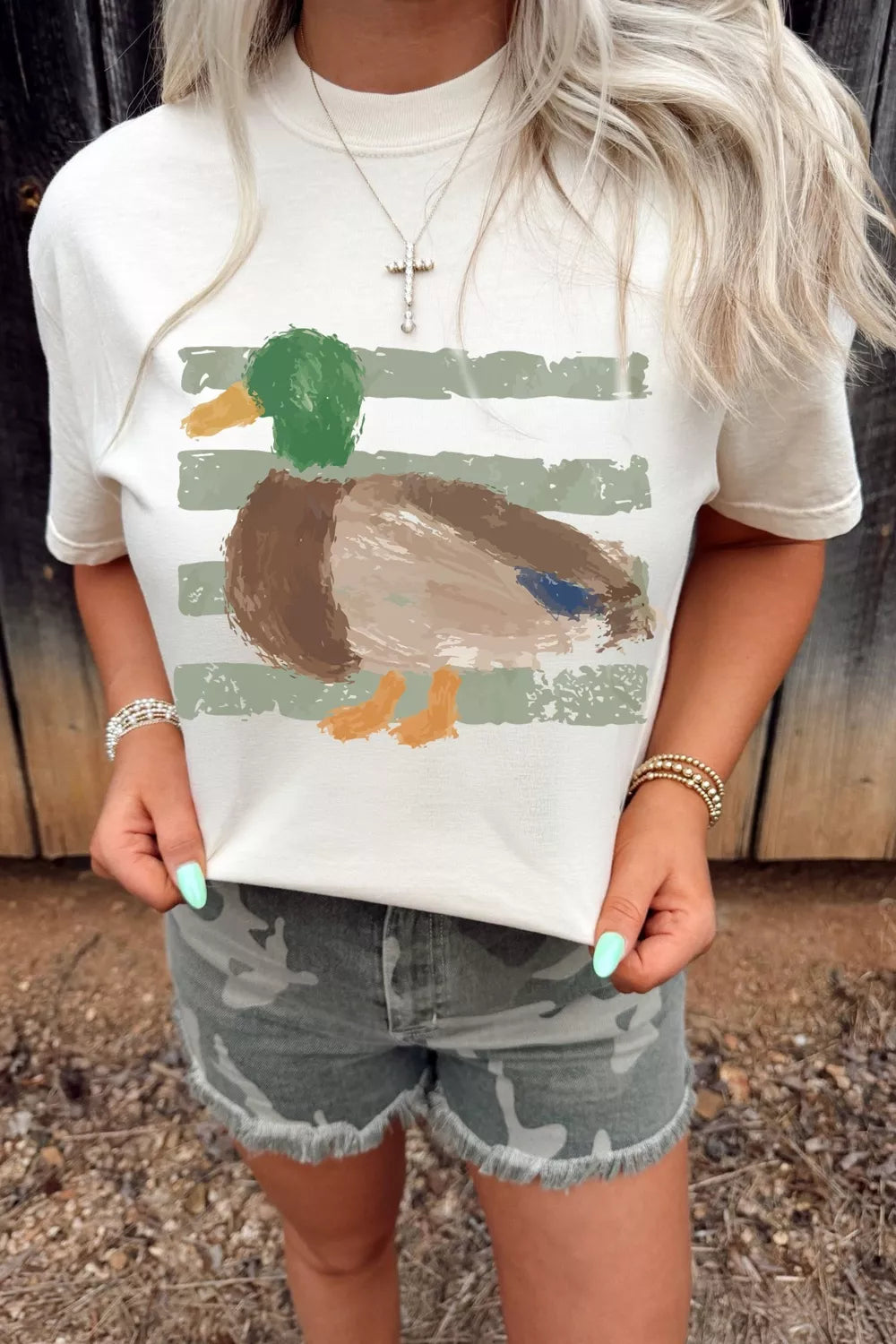 White short sleeve graphic tee with a painterly mallard duck design over muted green stripes, styled with camo shorts.