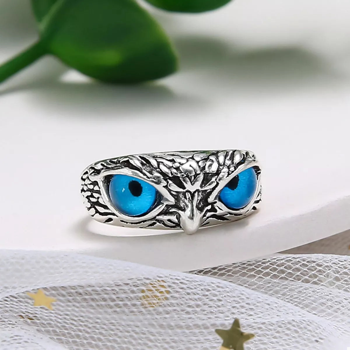 A silver-plated alloy ring with detailed owl features and vivid blue resin eyes, called the Nocturne Gaze Ring.