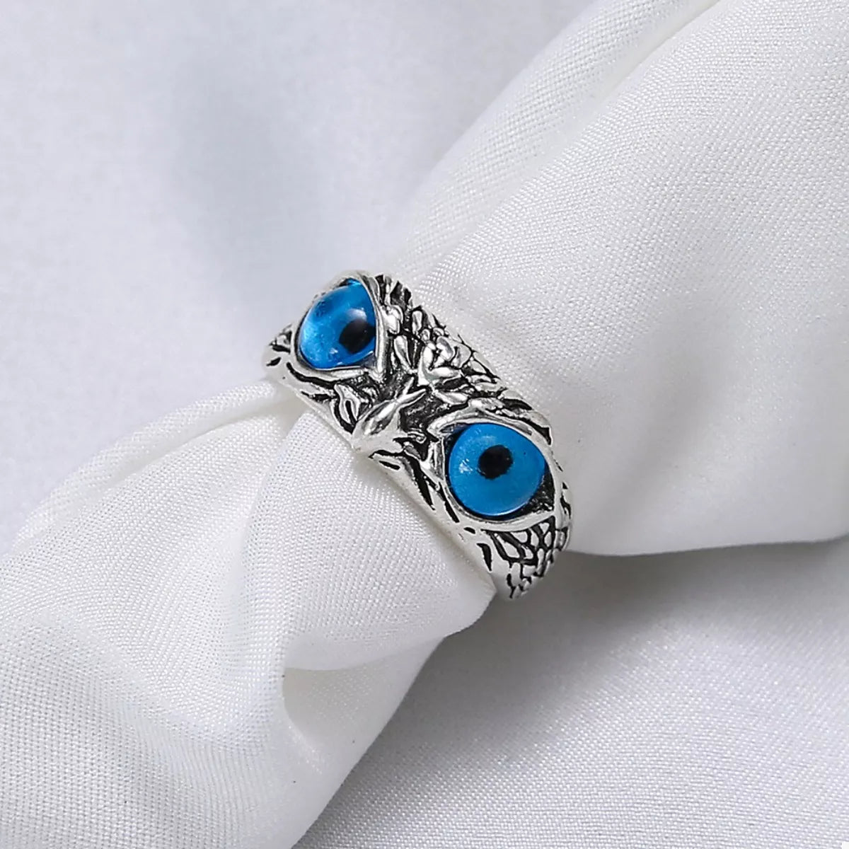 A silver-plated alloy ring with detailed owl features and vivid blue resin eyes, called the Nocturne Gaze Ring.
