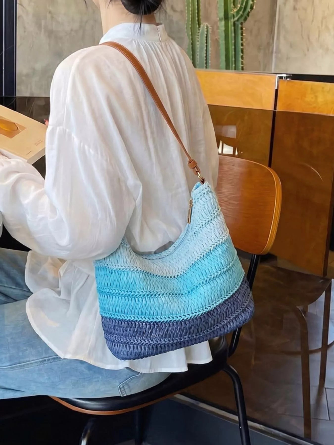 Medium woven straw shoulder bag with blue coastal stripes, curved silhouette, brown shoulder strap, and breezy summer styling.