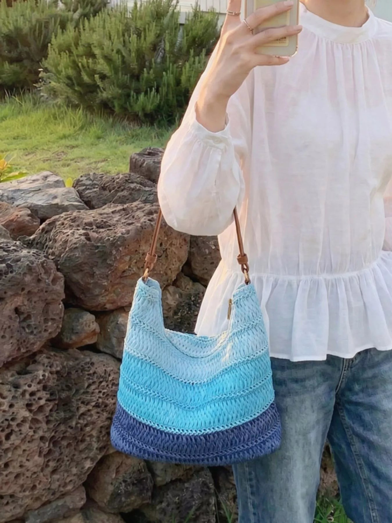 Medium woven straw shoulder bag with blue coastal stripes, curved silhouette, brown shoulder strap, and breezy summer styling.